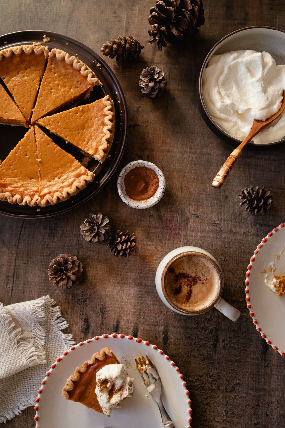 Overhead view of pumpkin pie with whipped cream and pinecone decorations on rustic wooden table.