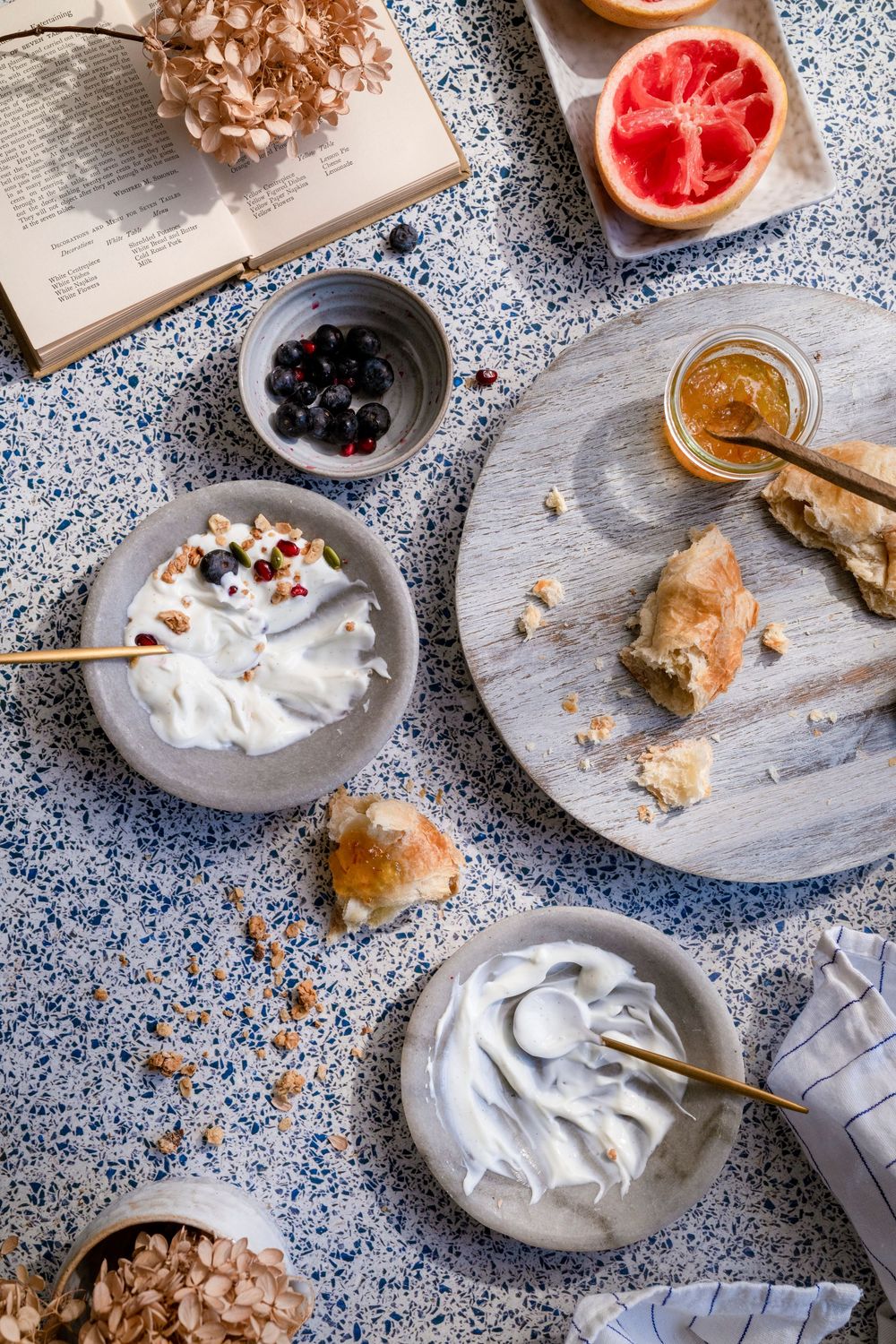 Breakfast spread with grapefruit, berries, yogurt and pastries on blue speckled surface.