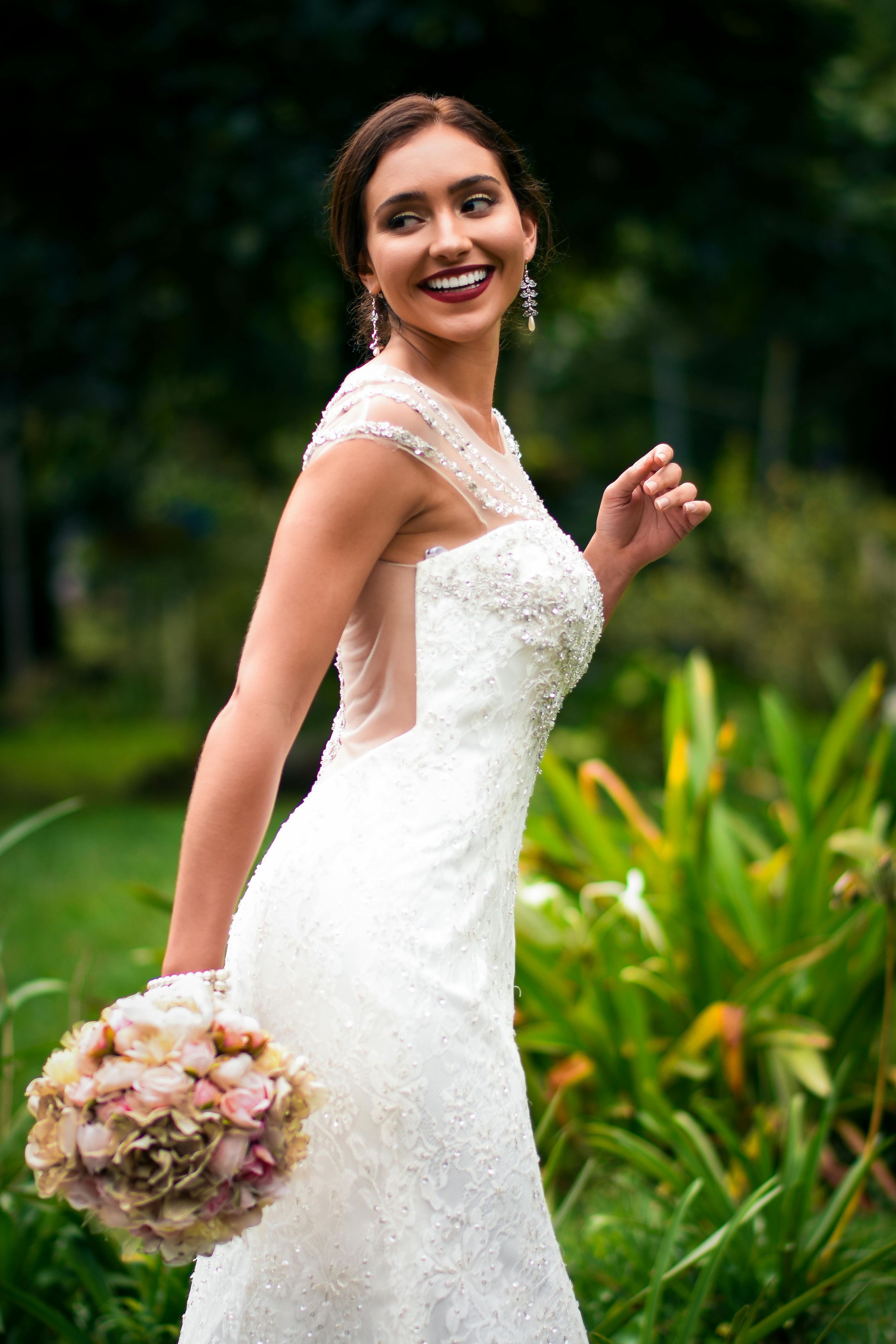 A bride in a white lace wedding dress smiles joyfully while holding a pink and white floral bouquet in a garden setting.