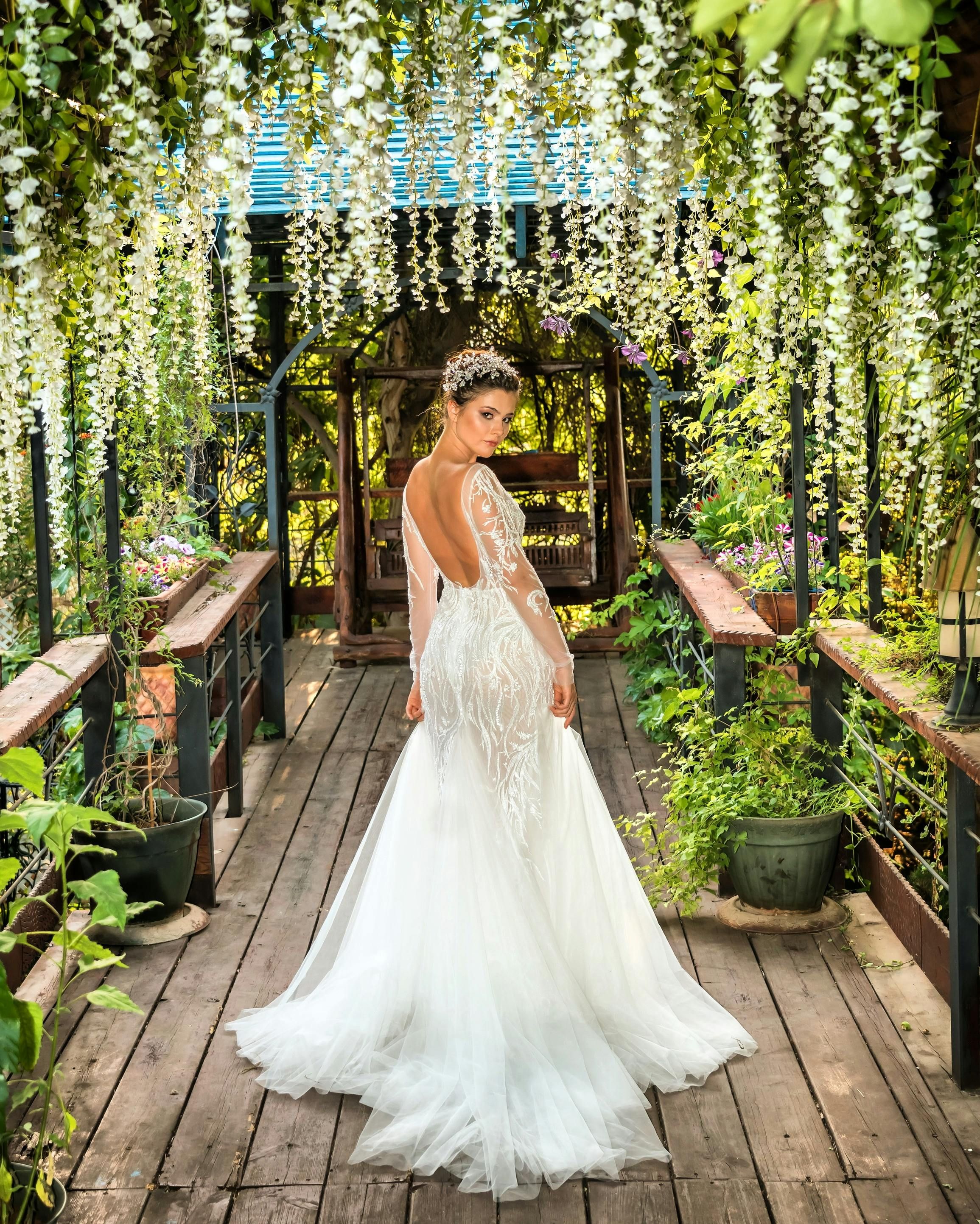 A stunning white wedding gown with open back design displayed in a garden pergola draped with cascading wisteria flowers.