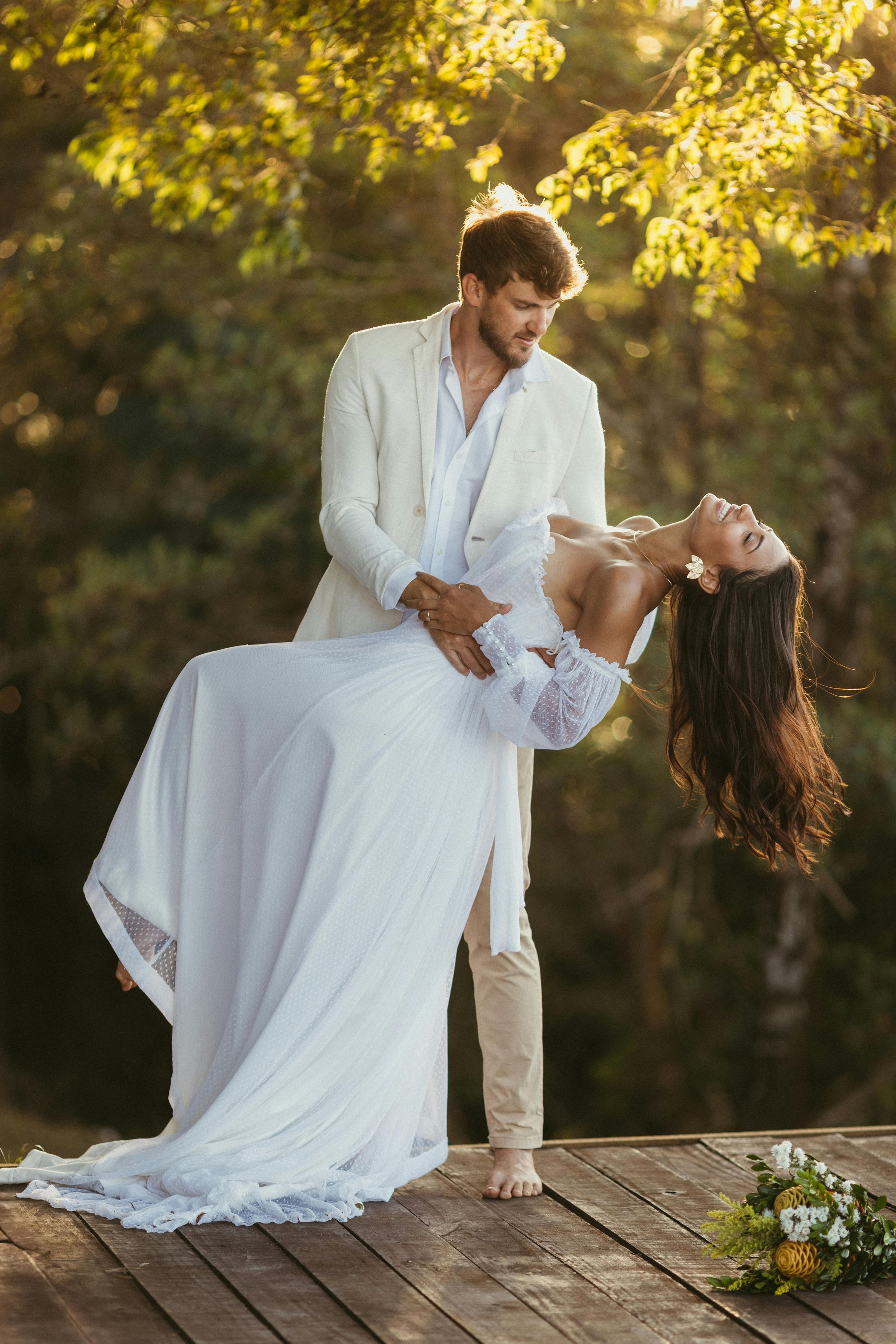 A couple in white attire dances romantically on a wooden deck during golden hour with autumn foliage in background.