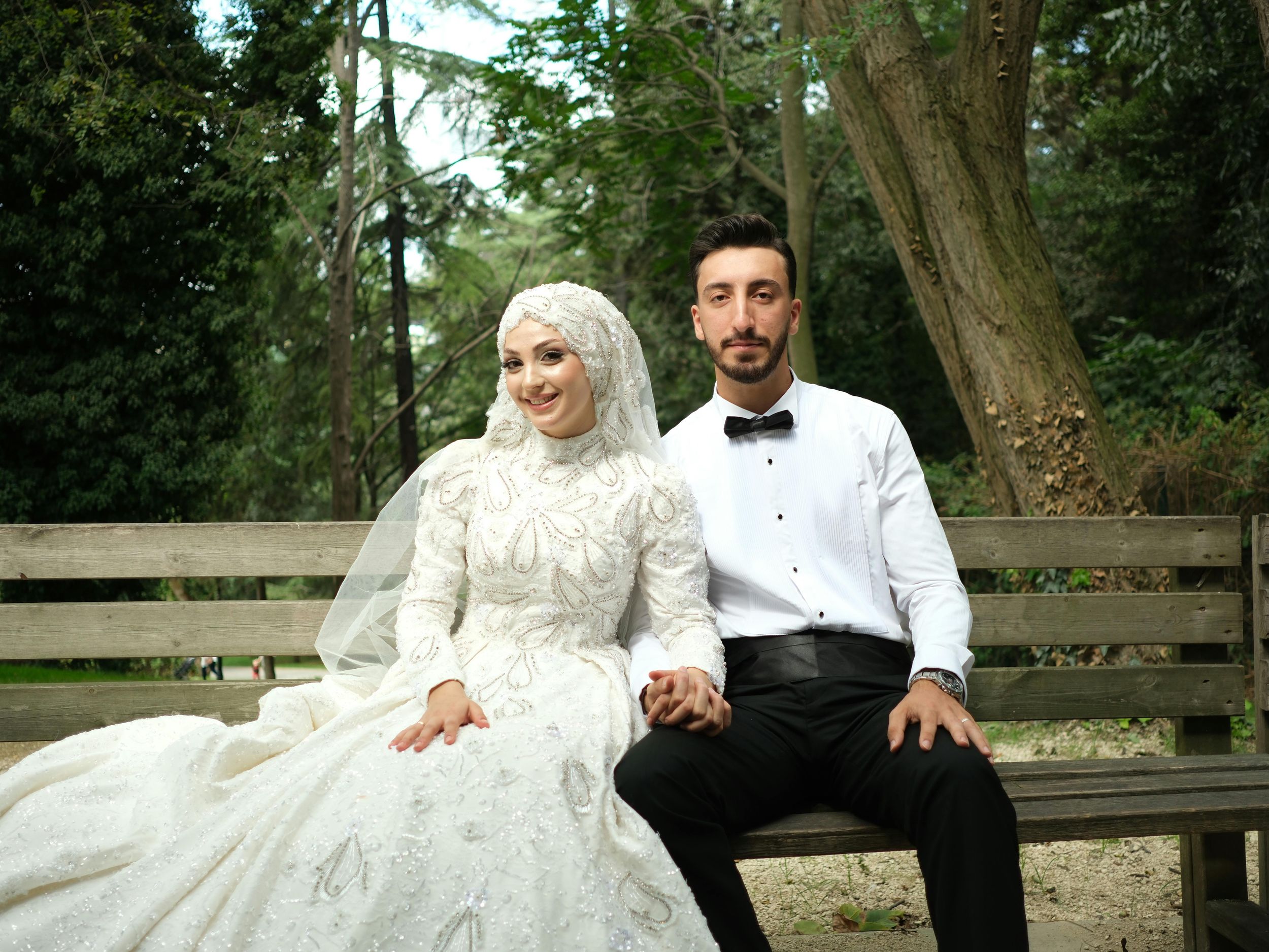 A wedding couple sits together on a wooden bench in a park setting surrounded by lush greenery.