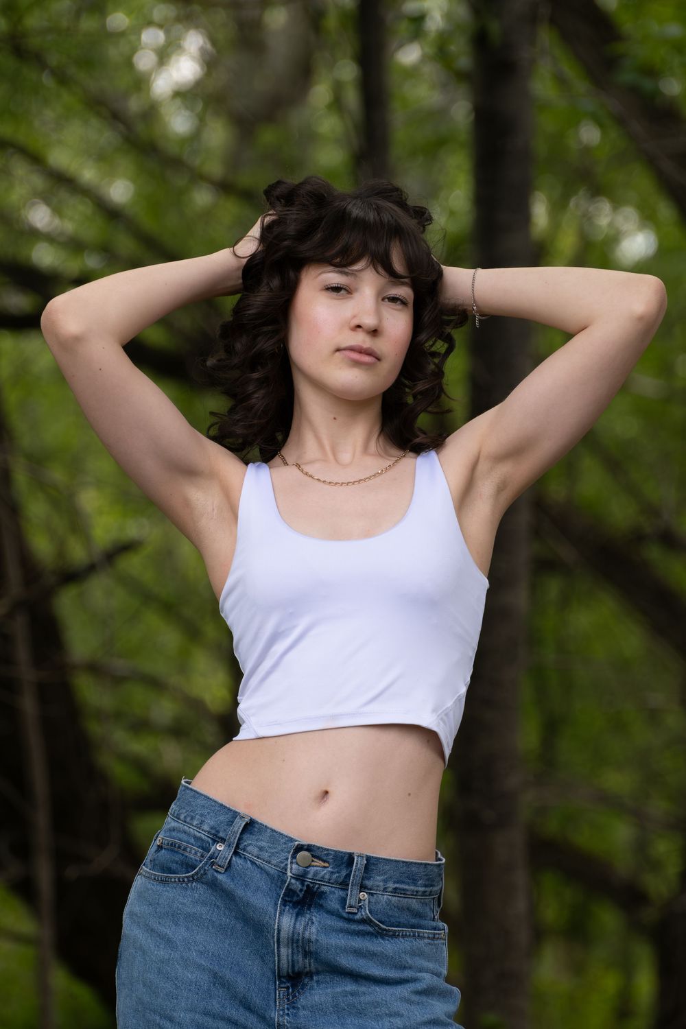 Young woman with curly dark hair in white crop top and jeans posing outdoors in a green forest setting.
