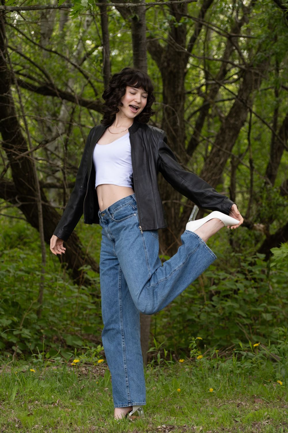 Young woman in jeans and leather jacket balancing on one leg outdoors in a lush green forest setting.