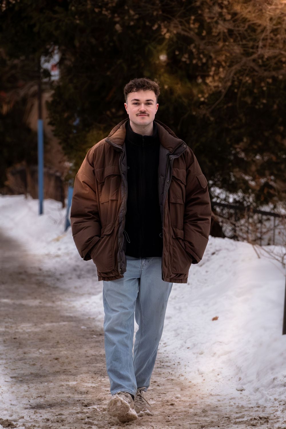 Person in brown winter jacket and light jeans walks on snowy path through winter landscape.