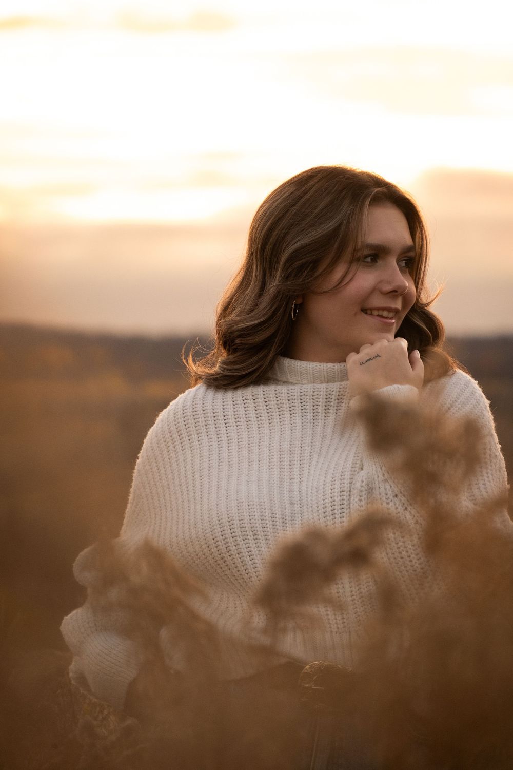 Person in white sweater smiling during golden hour sunset in a dreamy outdoor setting.