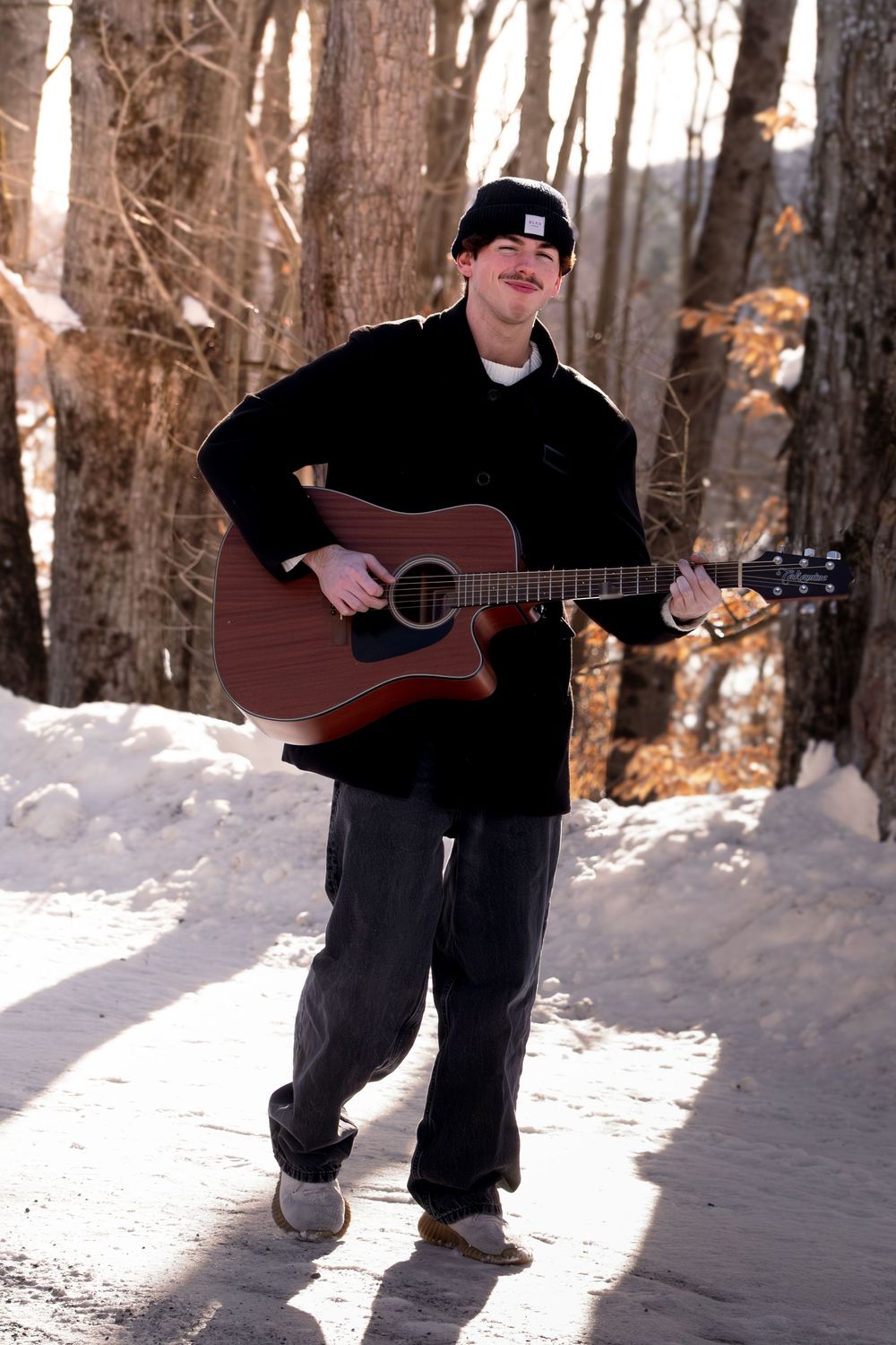 A musician plays acoustic guitar while sitting in snowy winter woods with sunlight filtering through bare trees.