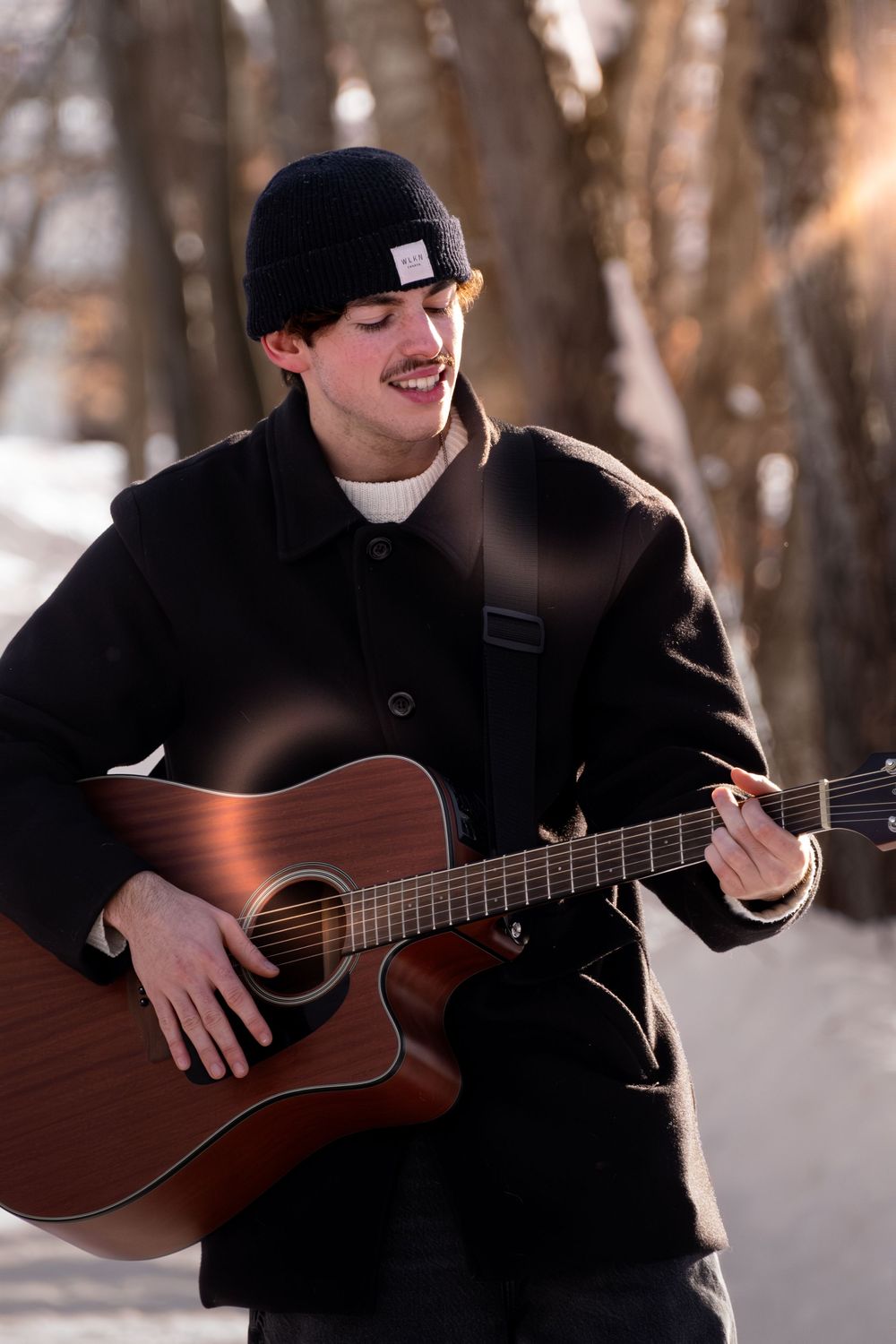 A person in black winter clothing plays an acoustic guitar outdoors in a snowy winter forest setting.