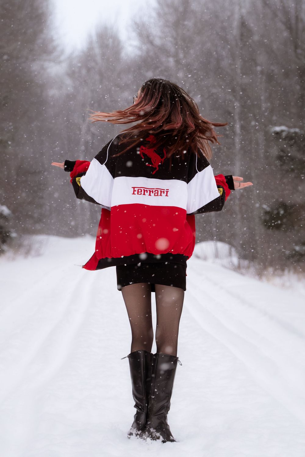 Person wearing a Ferrari jacket and boots dances joyfully in the snow on a winter forest path.