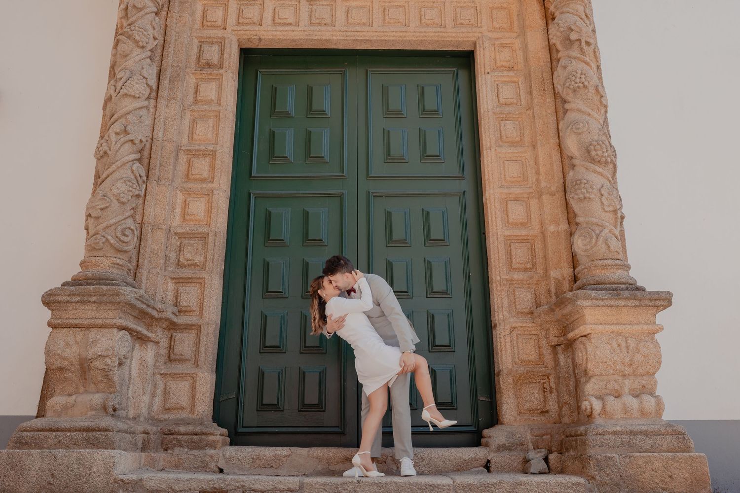 A couple embraces in front of large green wooden doors at an ornate stone church entrance.