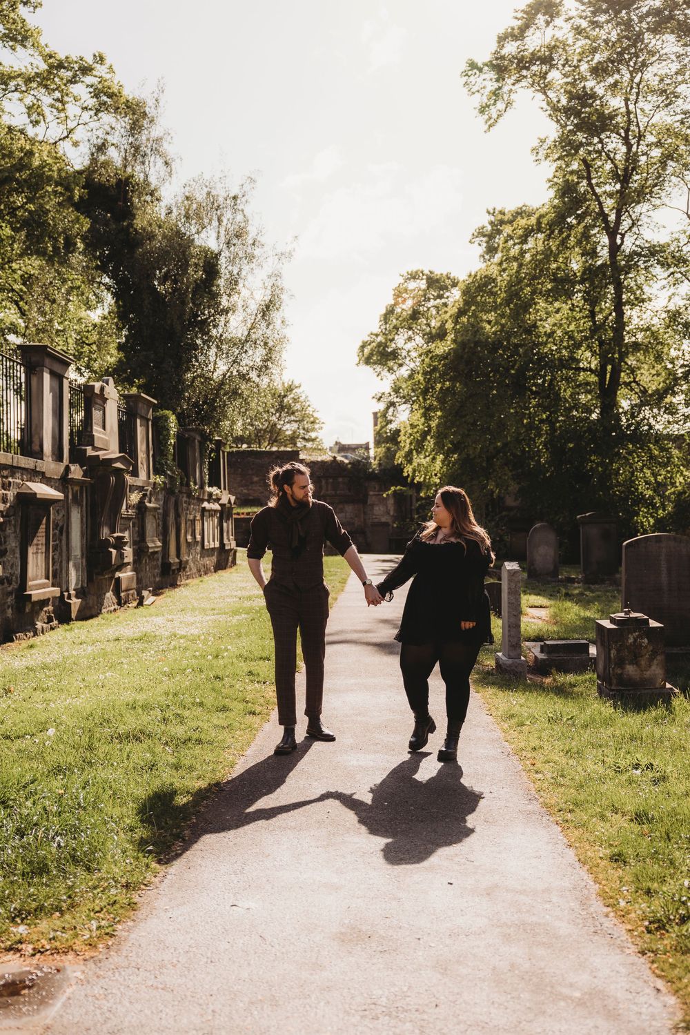 Two figures walk along a sunlit cemetery path between gravestones on a warm afternoon.