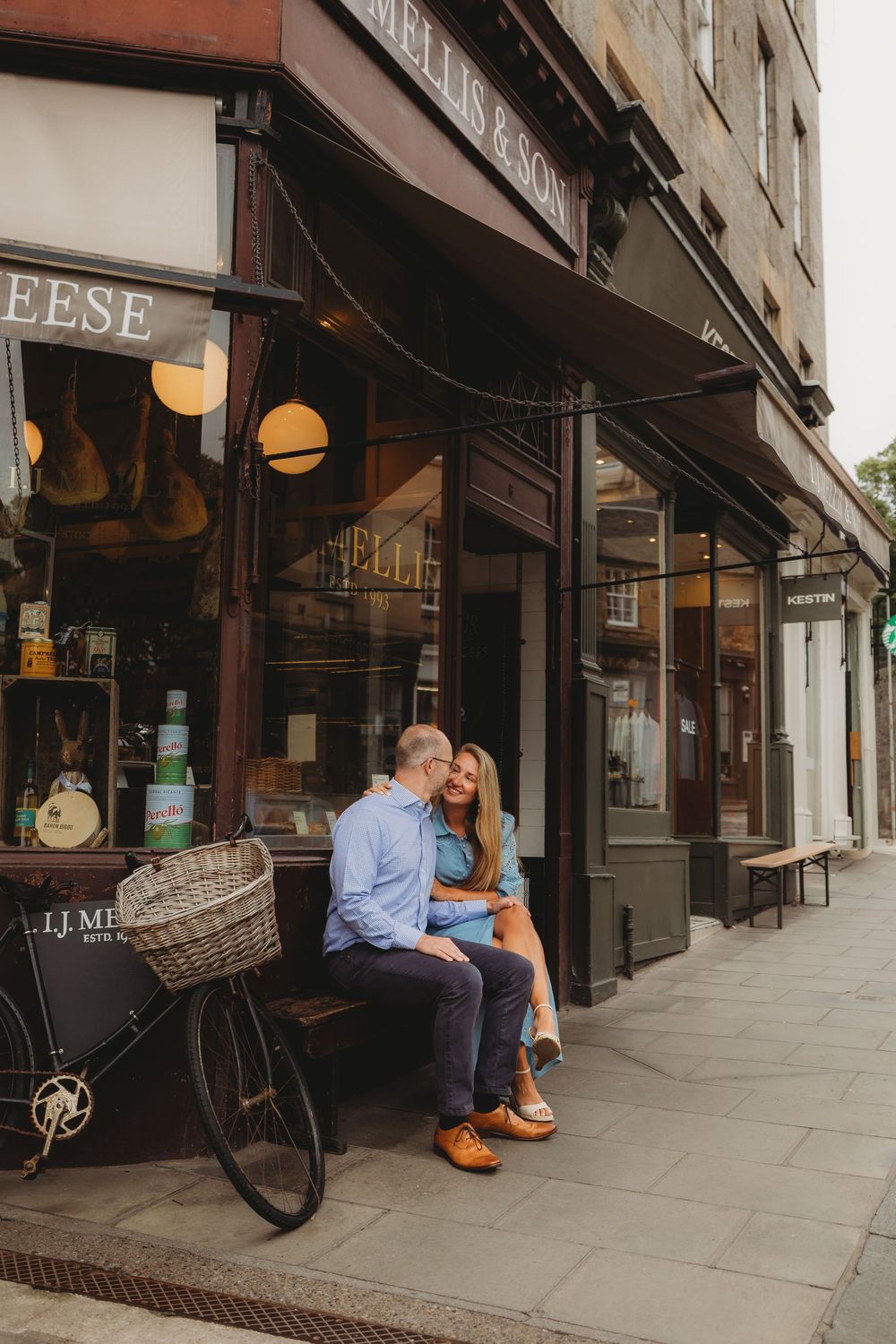 Couple sitting together on a bench outside storefronts with vintage bicycle and street lamps.