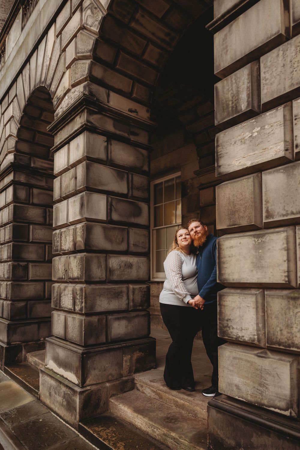 A romantic couple shares intimate moments in a historic stone archway corridor with dramatic lighting and shadows.