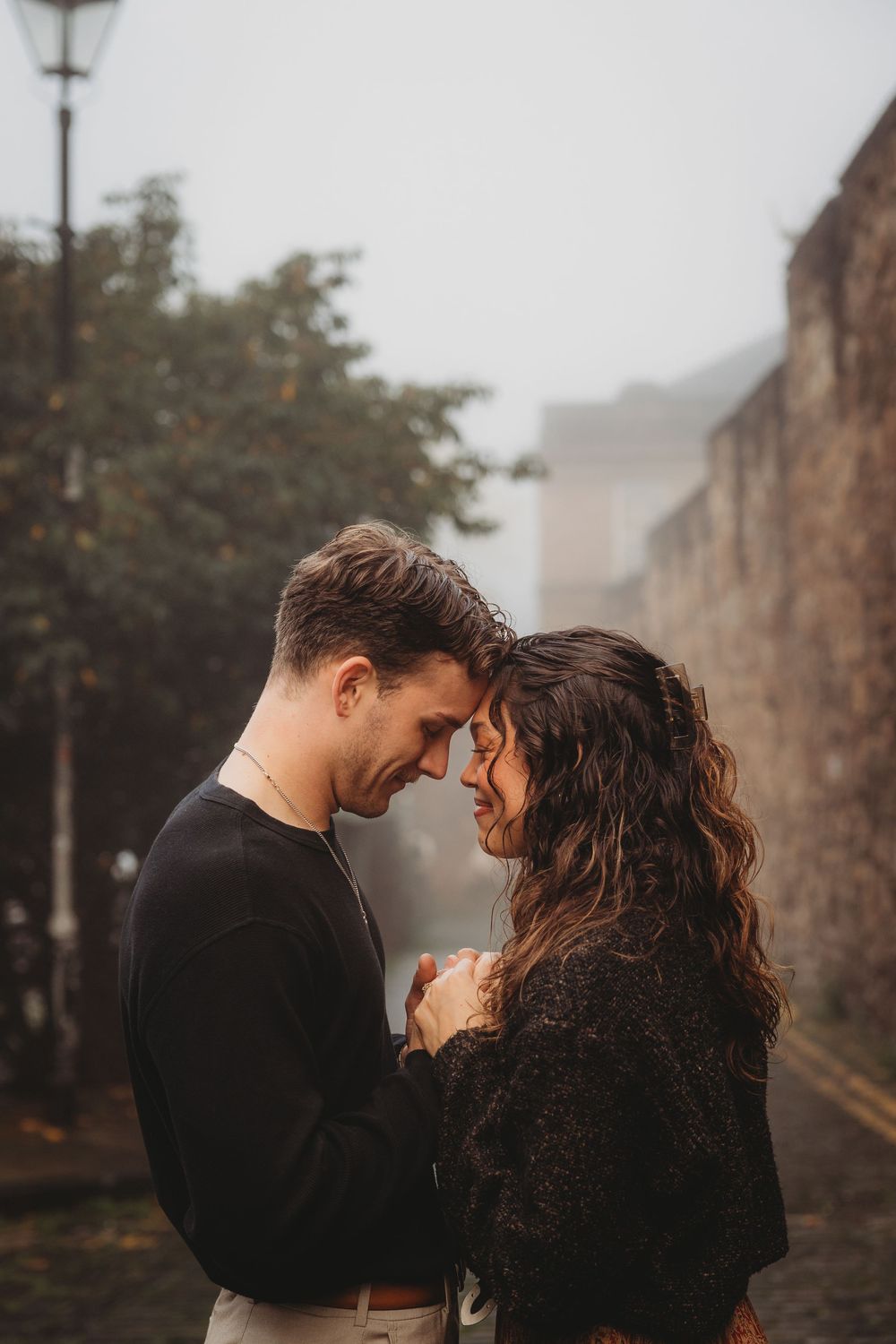 A romantic couple shares an intimate moment together on a misty autumn day against stone walls.