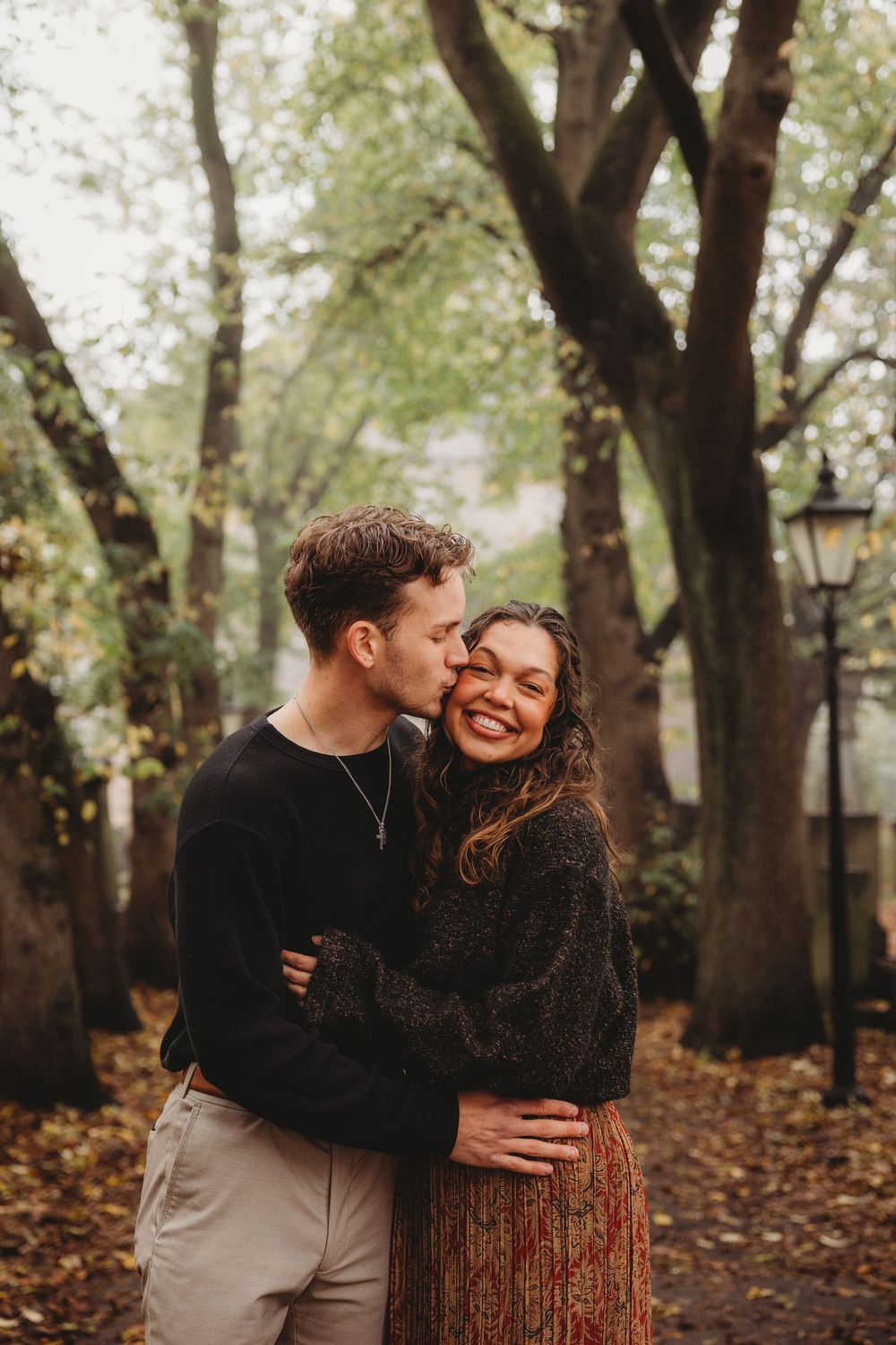 A couple embraces and laughs together among fall trees with scattered leaves on the ground.