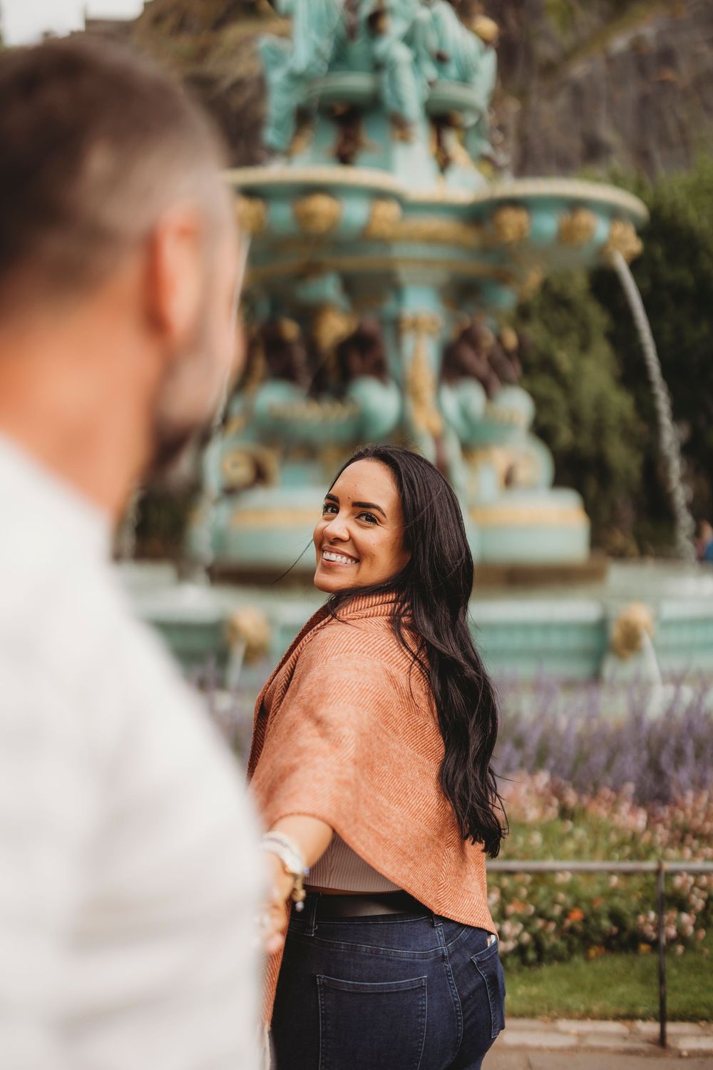 A couple shares a candid moment in front of an ornate turquoise fountain in a garden setting.