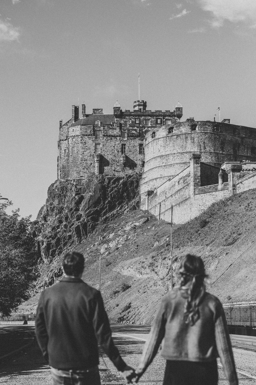A couple holds hands while looking up at the historic Edinburgh Castle perched on a rocky cliff in black and white.