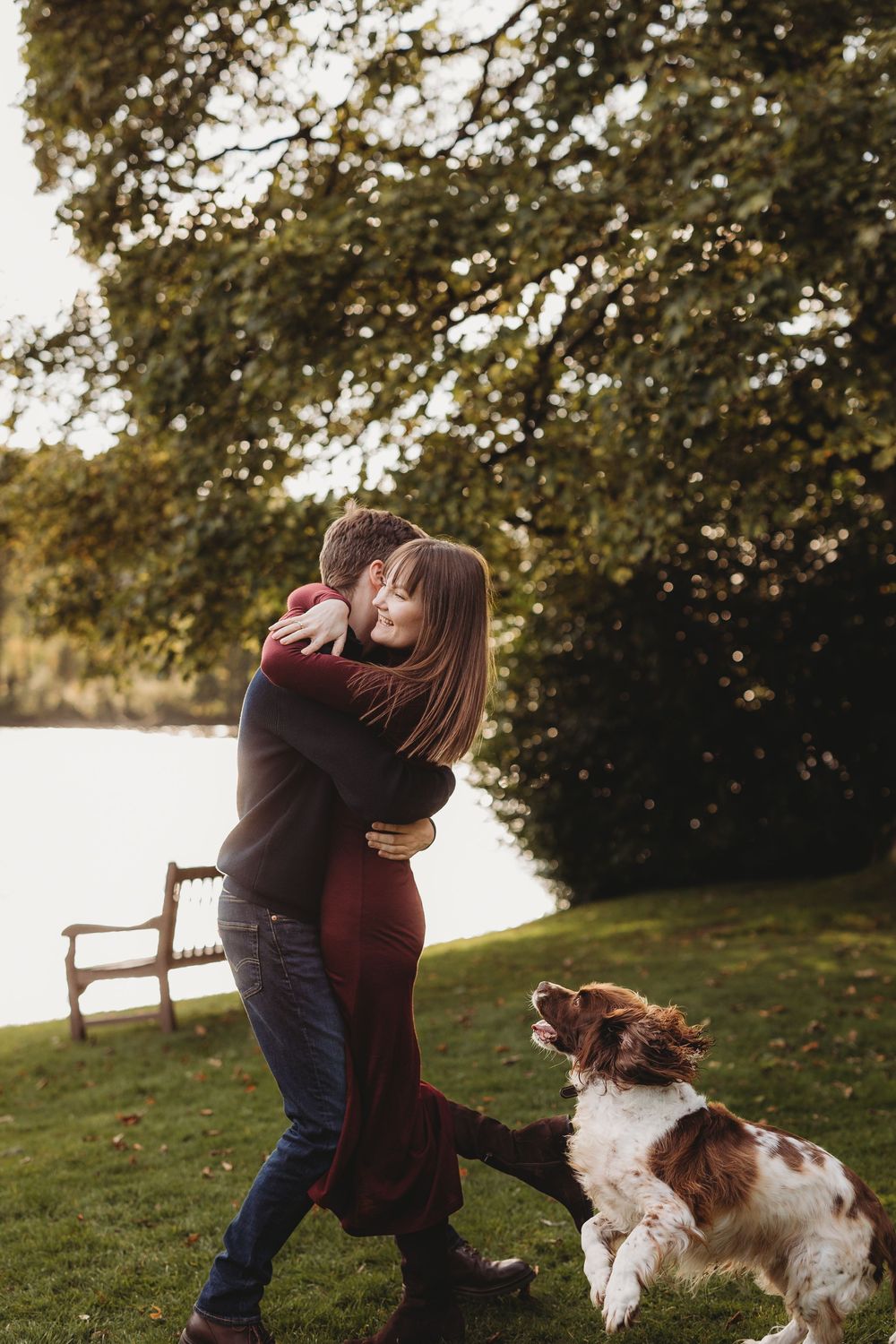 A couple embraces outdoors on a grassy lawn at sunset with their spaniel dog watching beside them.