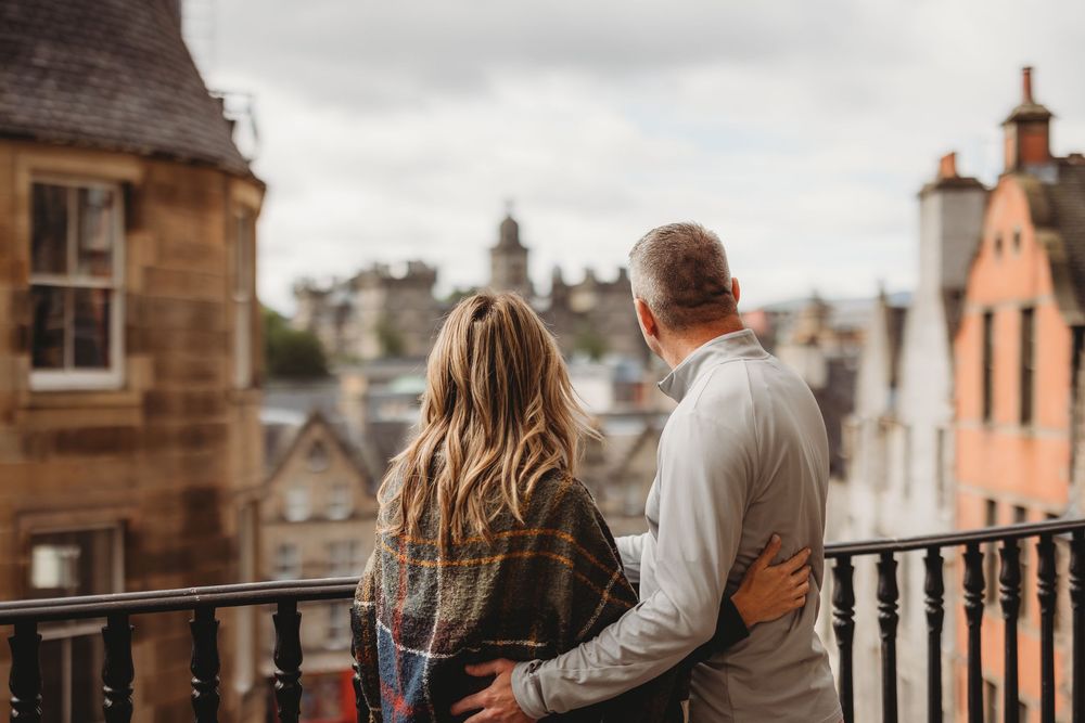 Couples enjoying scenic views from a historic stone balcony overlooking Edinburgh's historic cityscape on a cloudy day.