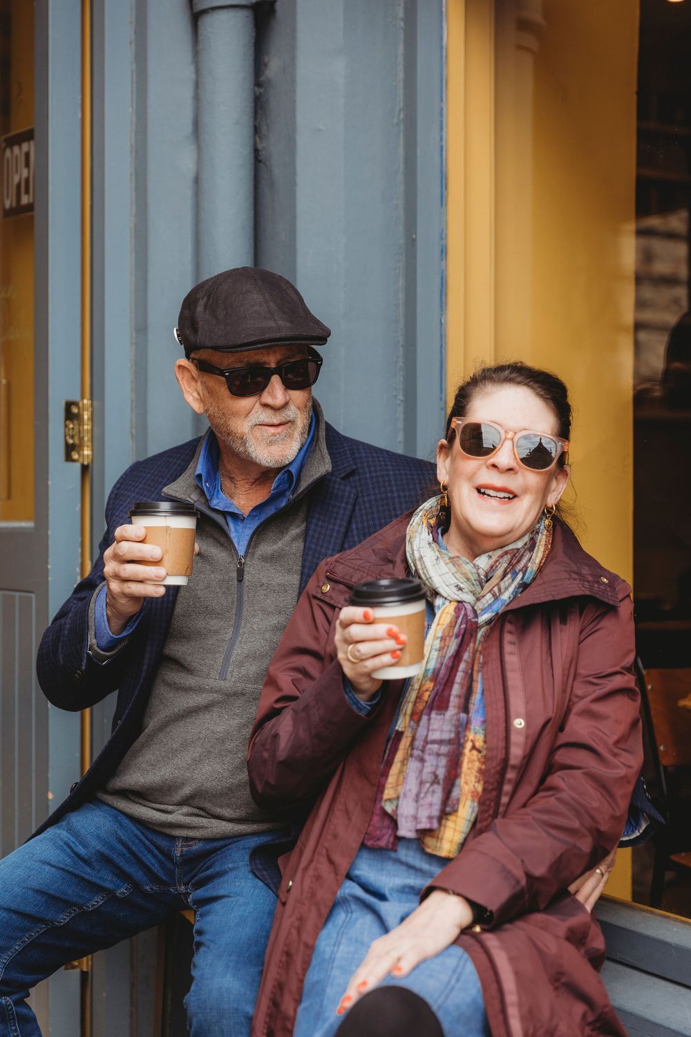 Two elderly friends share drinks and laughter while sitting outside on a sunny day.