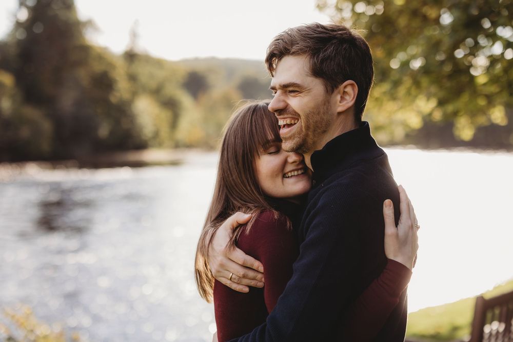 A romantic couple embraces and laughs together by a scenic waterfront during a golden sunset photoshoot.
