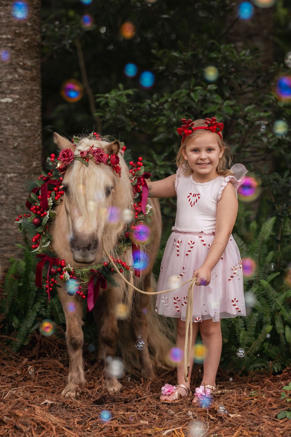 A pony adorned with red flowers stands next to a child in a white dress with red accents under colorful bokeh lights.