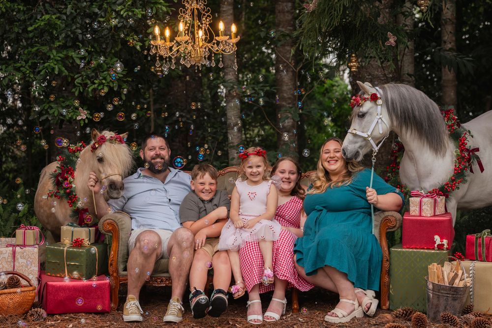 A group sits together on a vintage couch with a decorated pony and holiday presents in an outdoor setting.