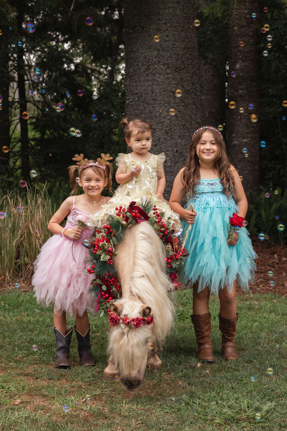 Three children in tulle dresses pose with a decorated pony amid floating bubbles in a forest setting.