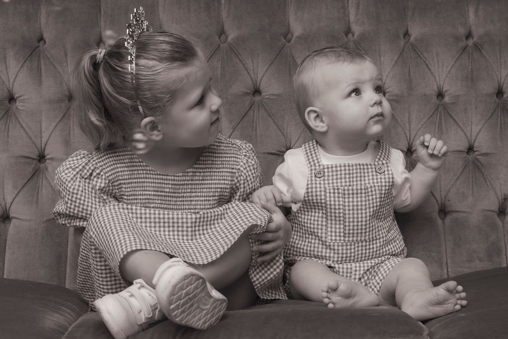 A black and white portrait of two people in gingham outfits sitting on a tufted couch.