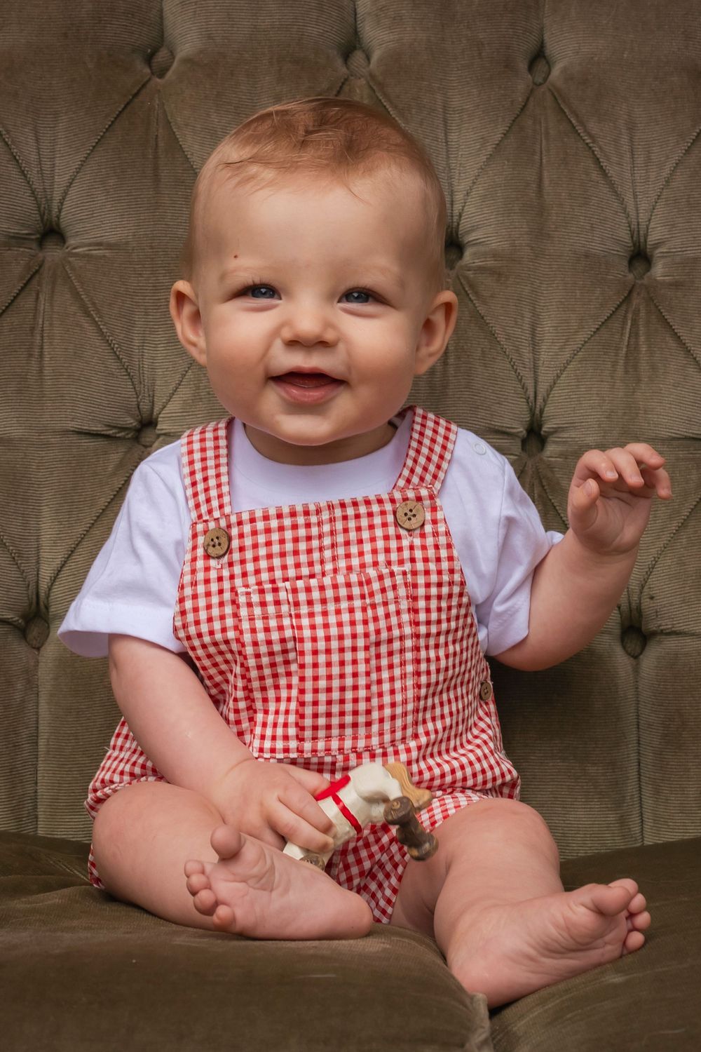 A baby wearing red and white gingham overalls sits smiling on a tufted velvet couch.