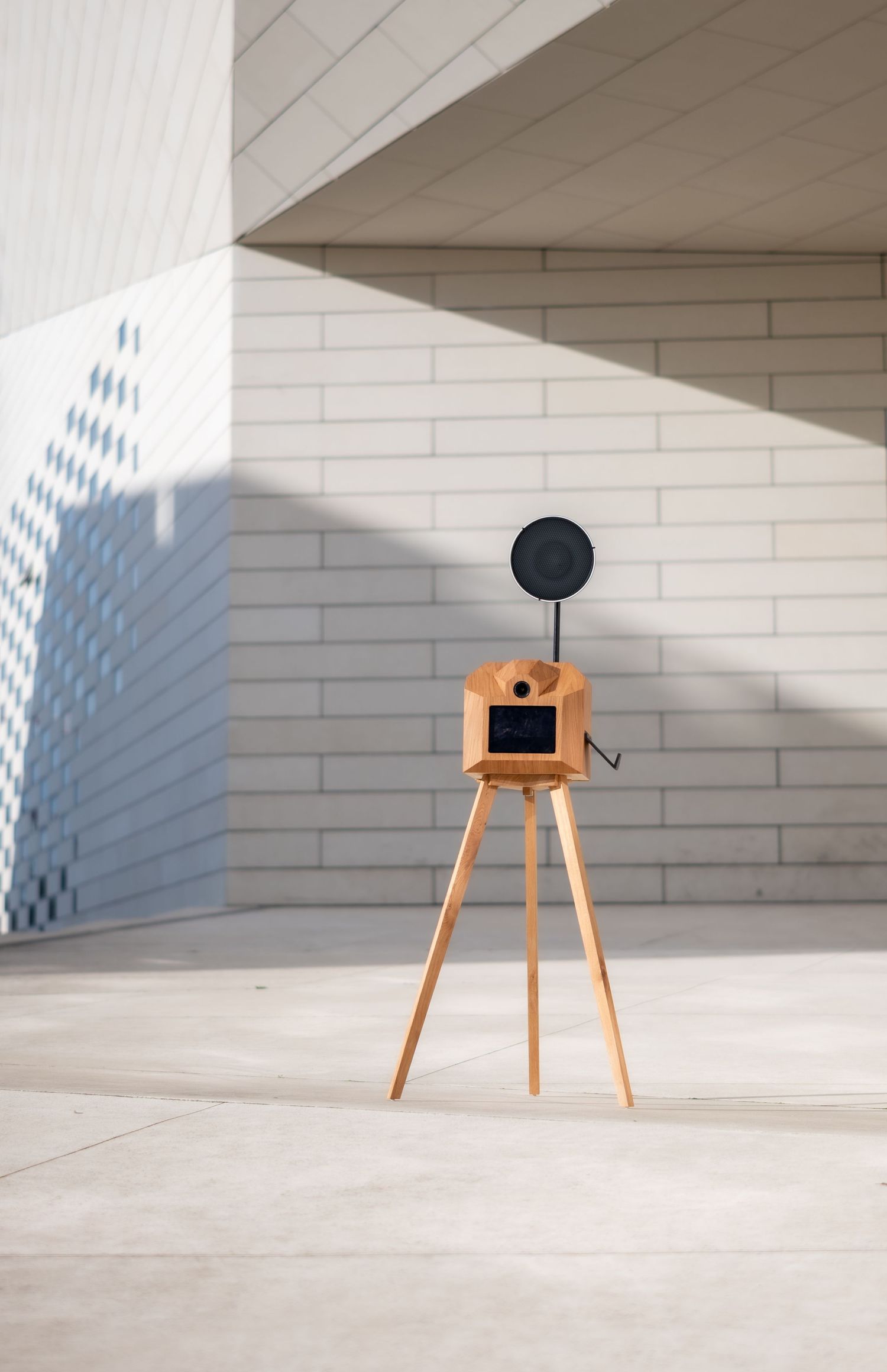 Minimalist wooden stool with black camera stands alone in bright white architectural space with dramatic shadows.