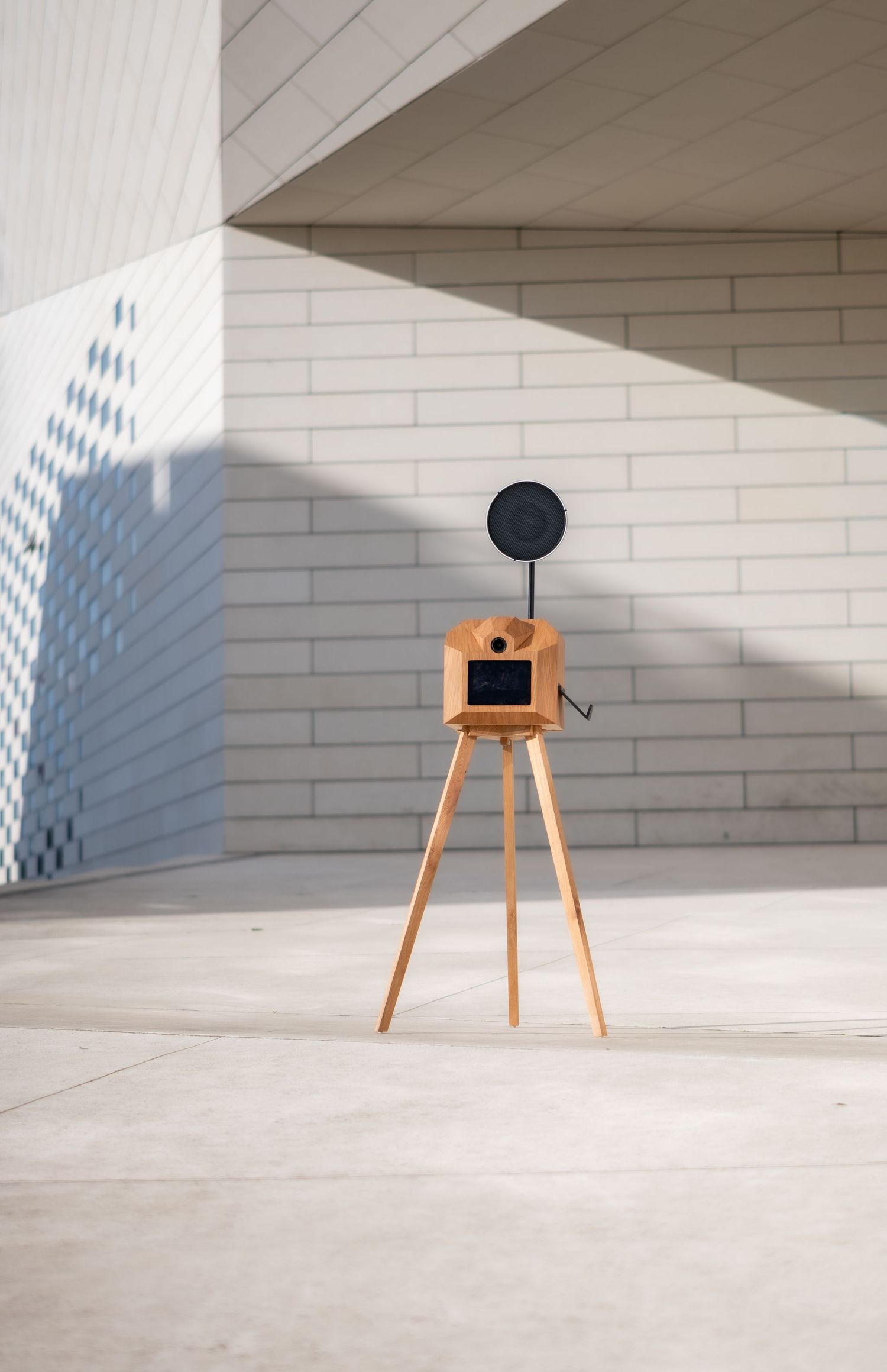 A black circular object on a wooden tripod stand creates shadows on white walls in a minimalist architectural space.