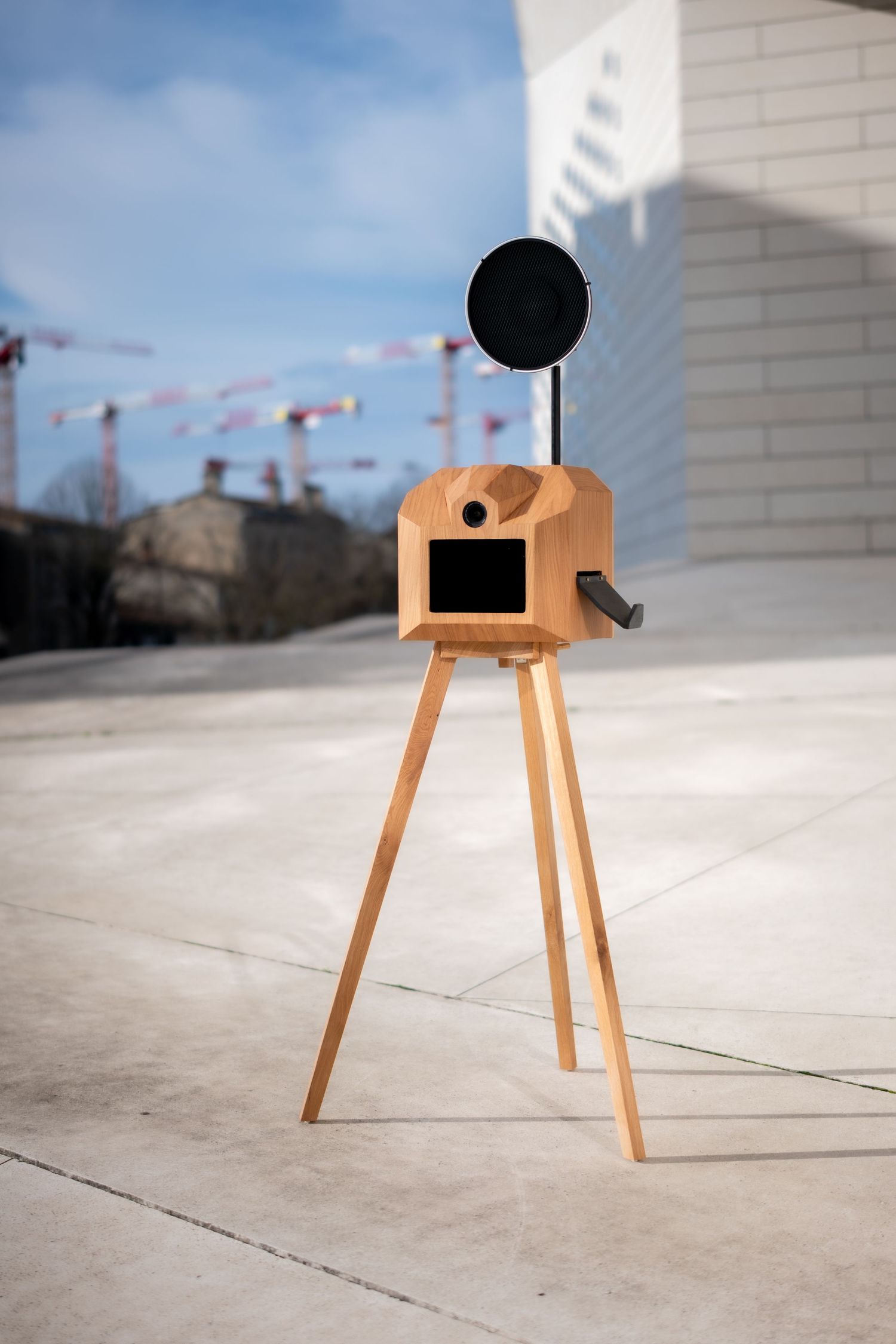 A minimalist wooden camera-shaped birdhouse with tripod legs sits on a concrete surface against a white wall.