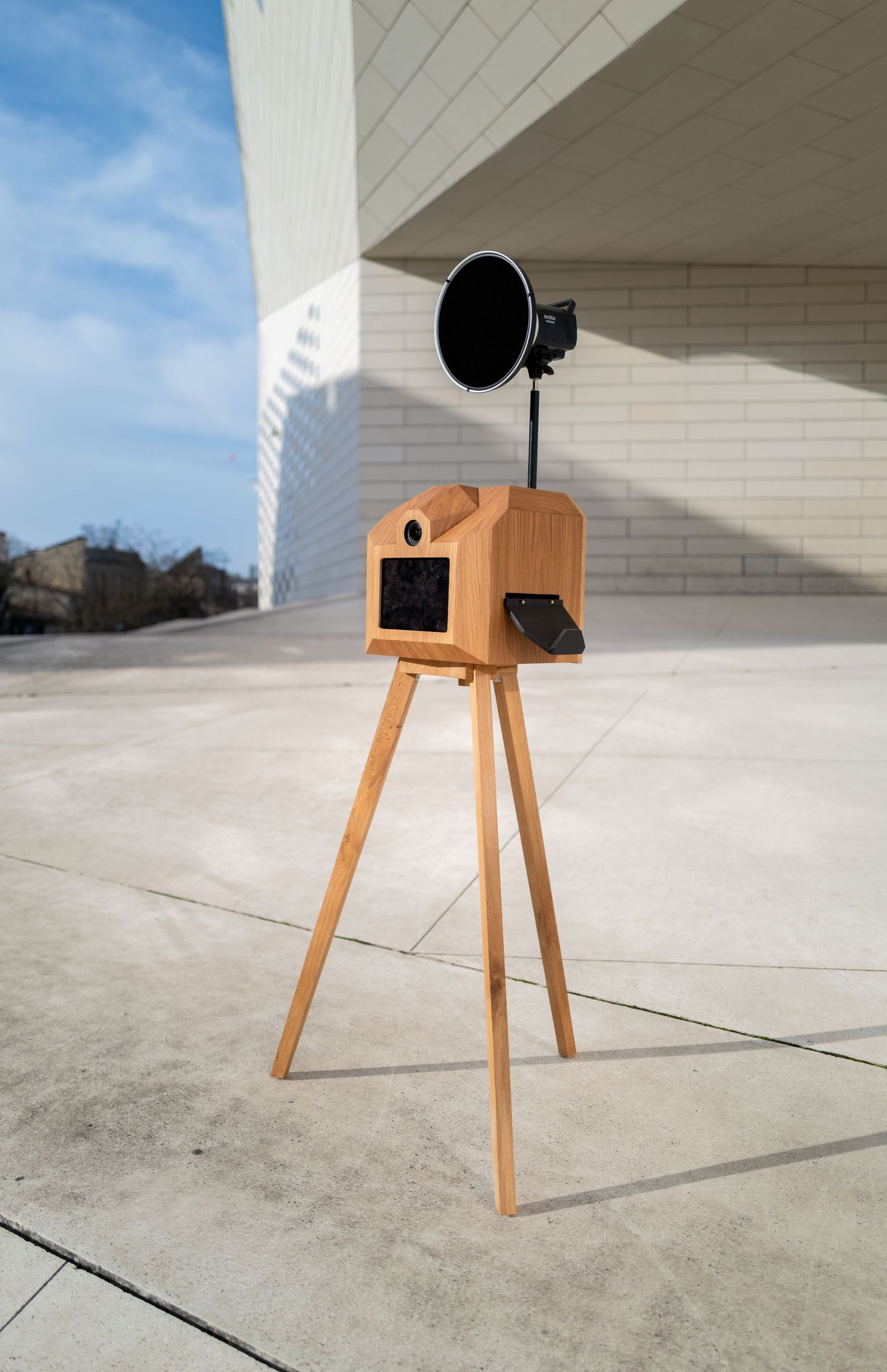 Wooden photo booth with tripod legs and black circular flash mounted on top stands against white building exterior.