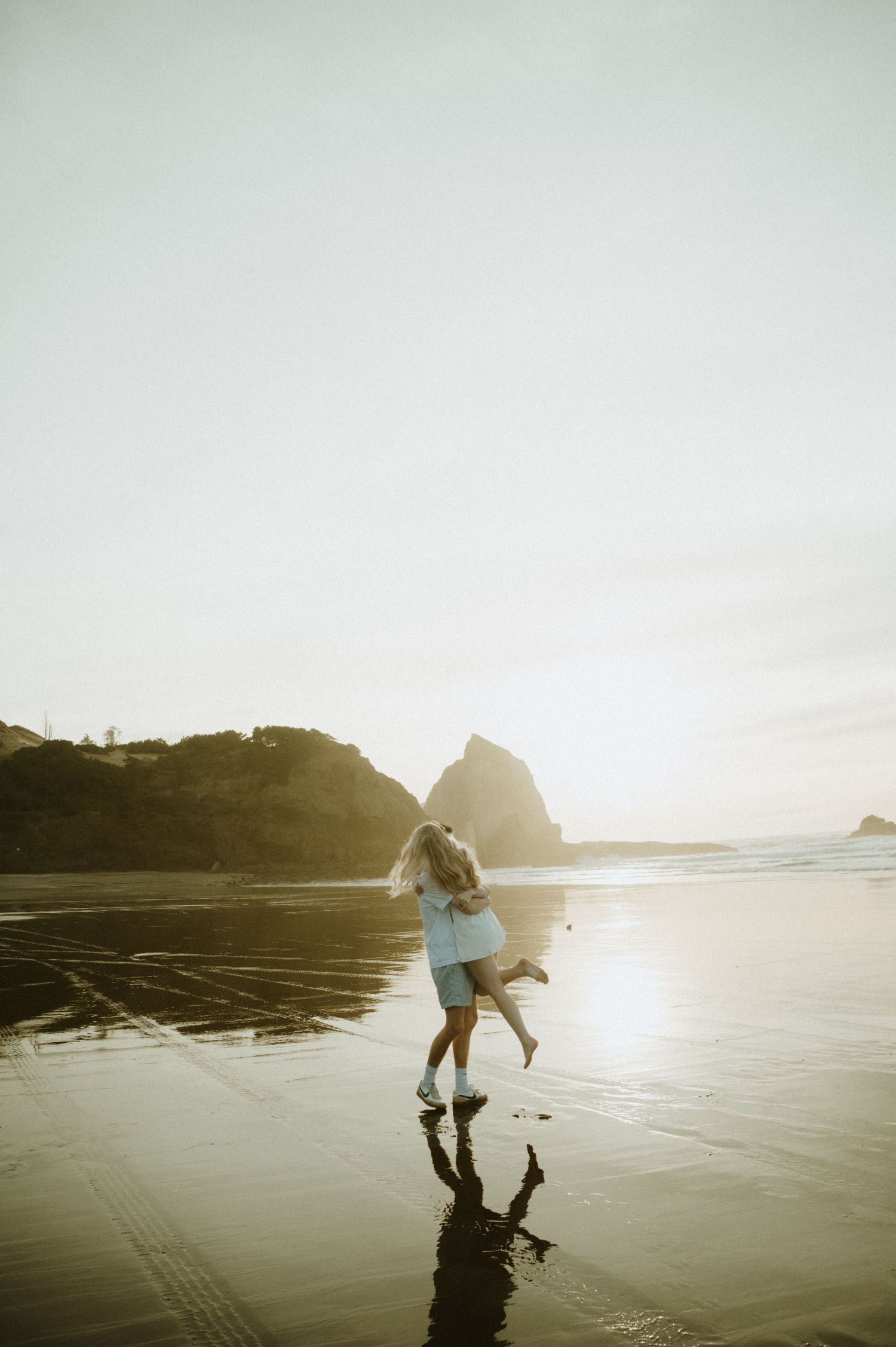 Couple playfully spins together on a sandy beach at sunset with coastal rocks in the distance.