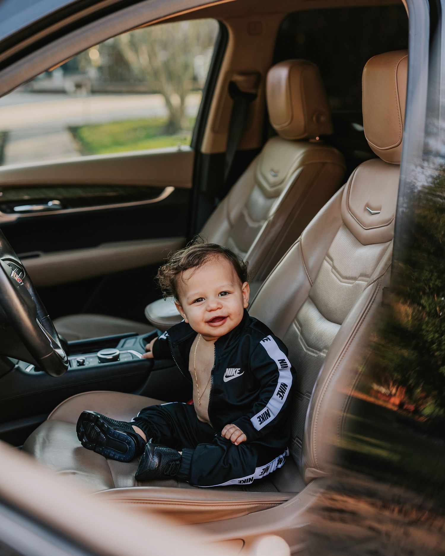 Happy toddler in Nike tracksuit sitting in luxury car driver's seat.