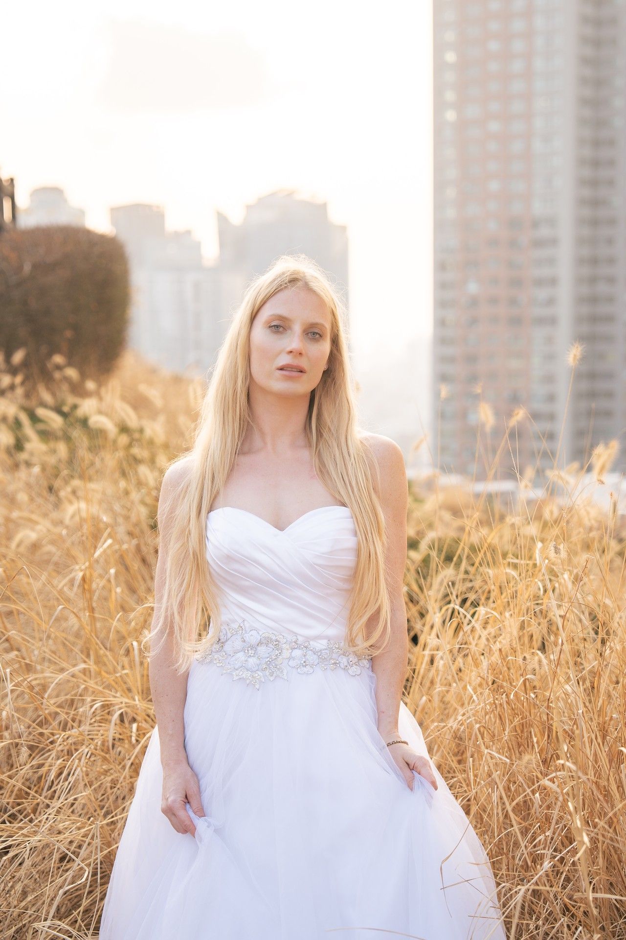 A bride in a white strapless wedding gown poses in tall golden grass with city buildings in the background at sunset.