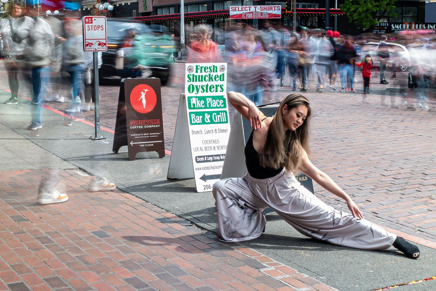 A dancer performs a graceful split pose on a busy brick sidewalk with blurred pedestrians moving in the background.