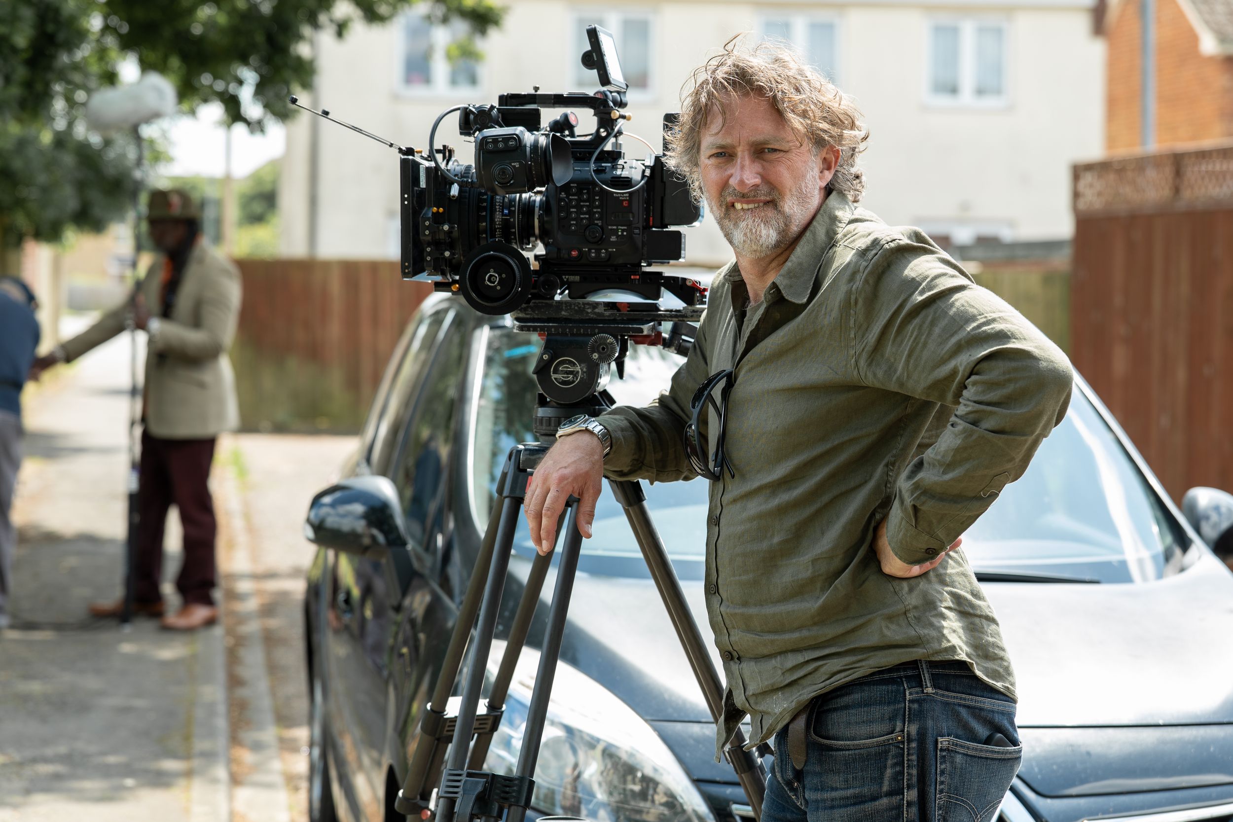 Camera operator with professional film equipment standing next to a car on a residential street.