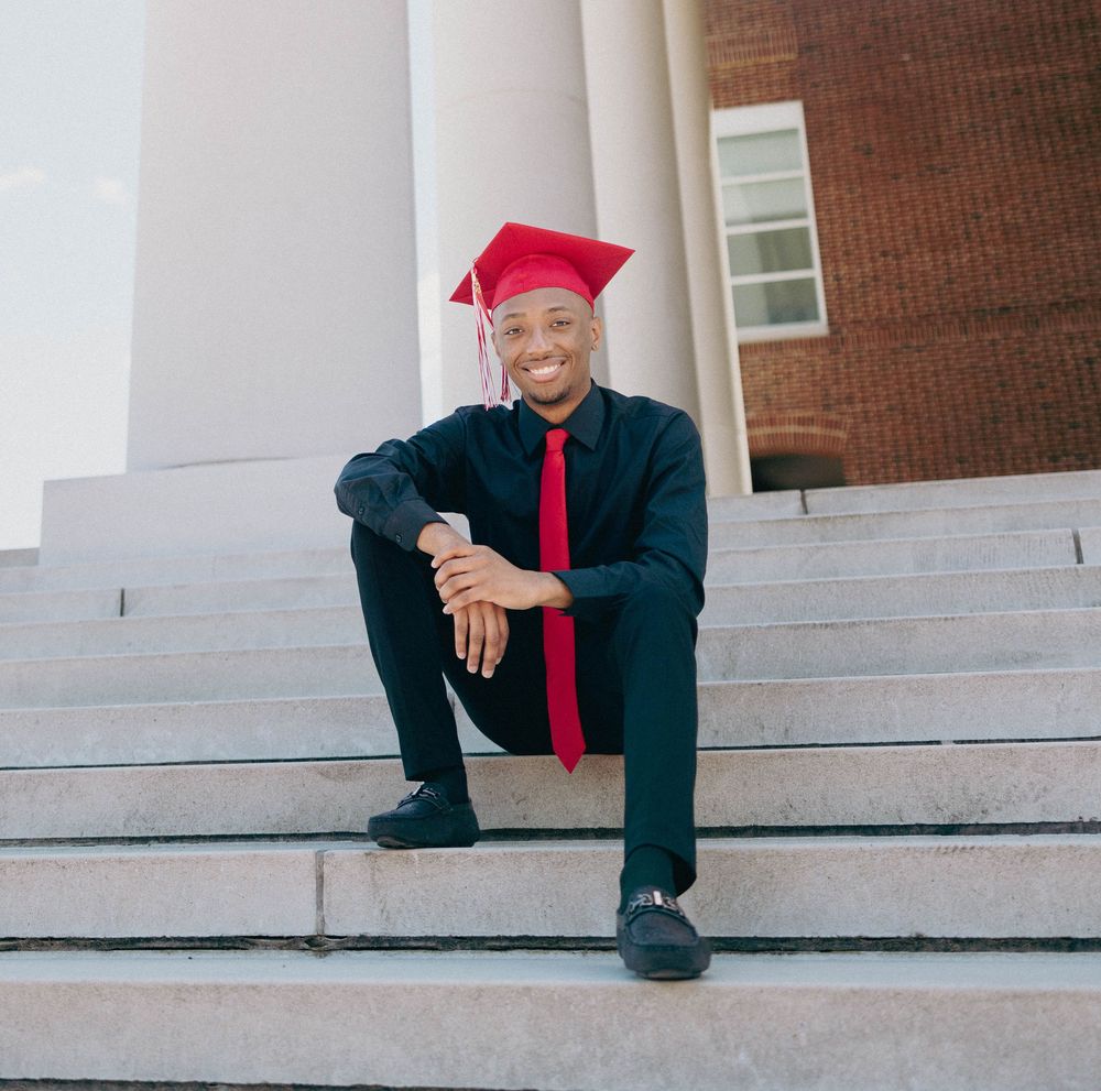 Graduate in black academic regalia sits on concrete steps during photo session.