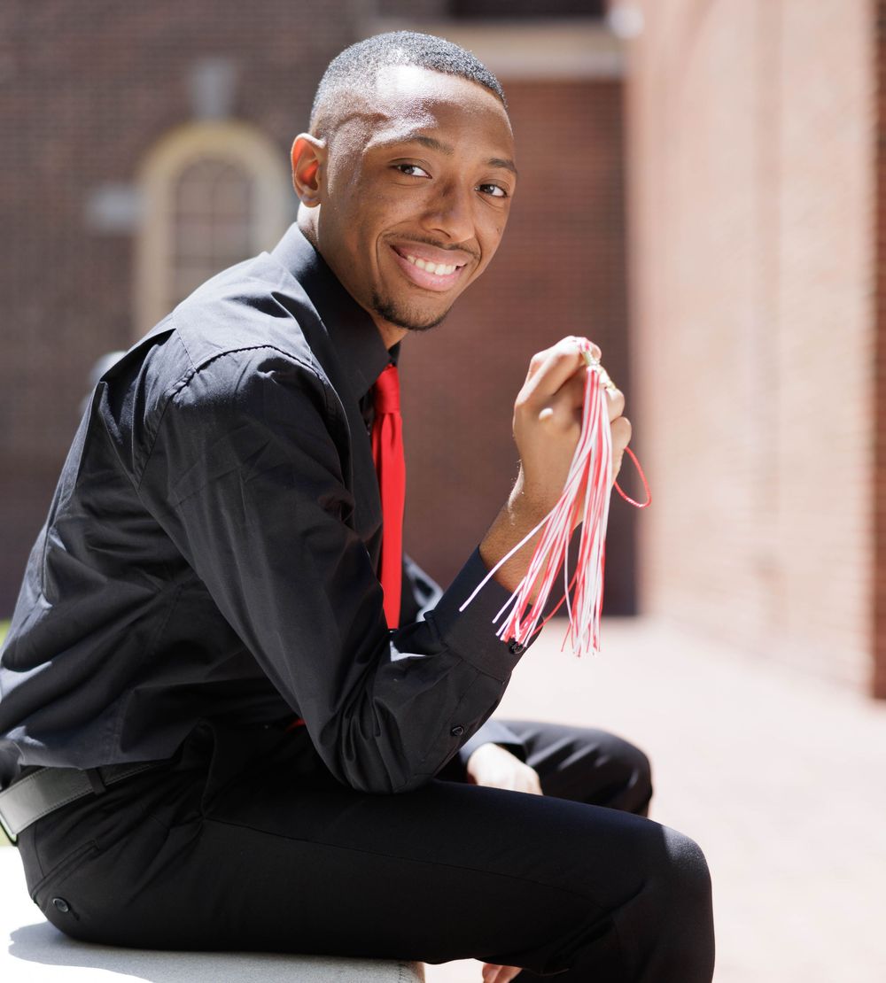 Graduate in black gown and red stole poses on campus steps.