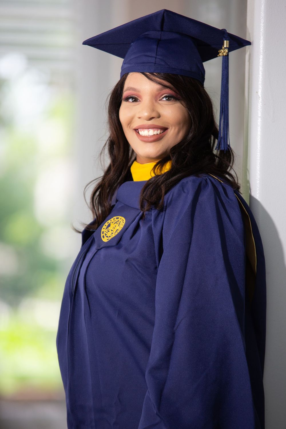 Graduate in navy blue academic regalia poses indoors near window.