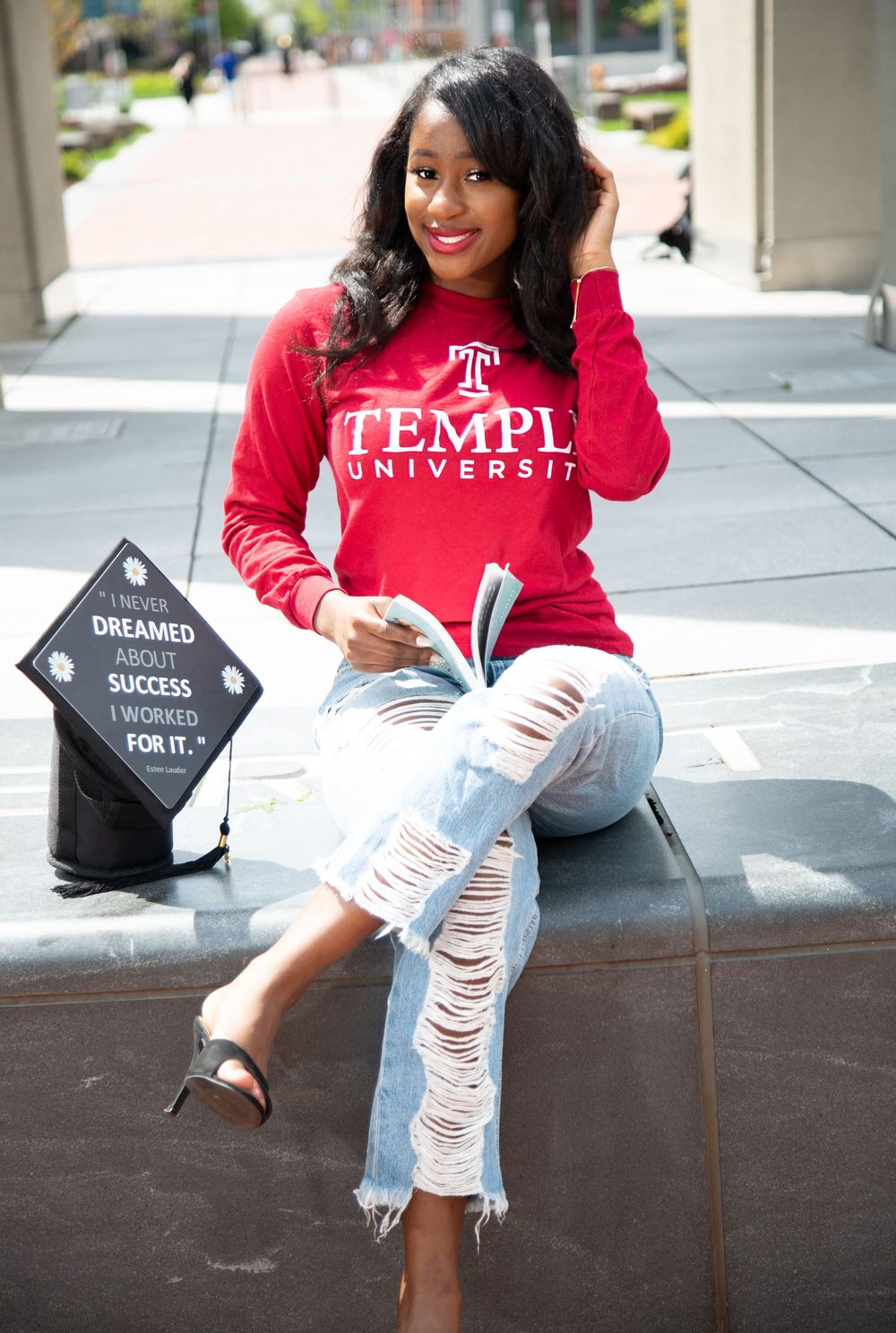 Graduate in red Temple University sweatshirt poses with ripped jeans and graduation cap signs for celebratory photos.