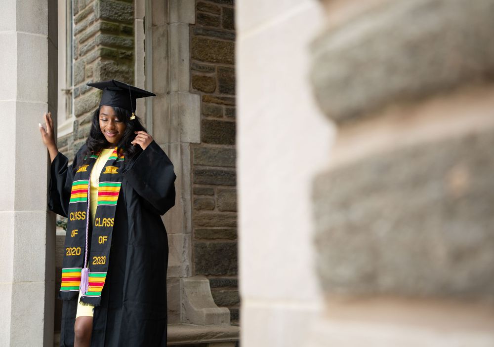 Graduate in traditional black gown and cultural stole poses between stone pillars.