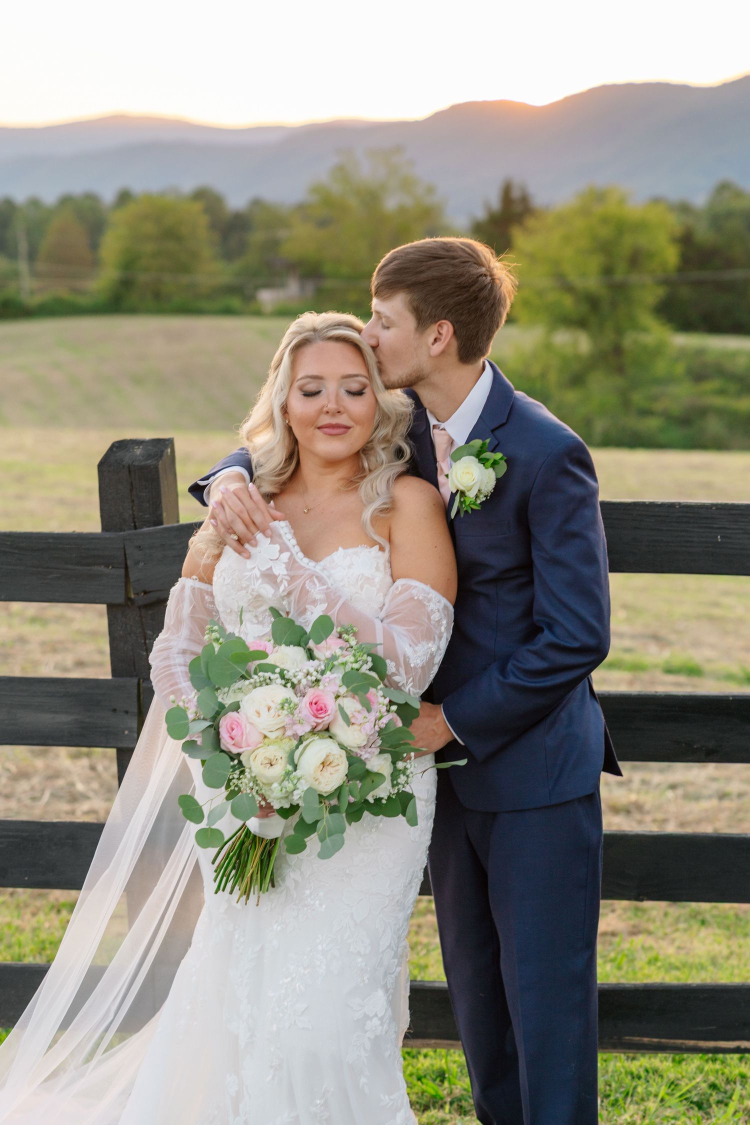 A romantic wedding couple embraces in front of a scenic mountain landscape with wooden fences and lush greenery.