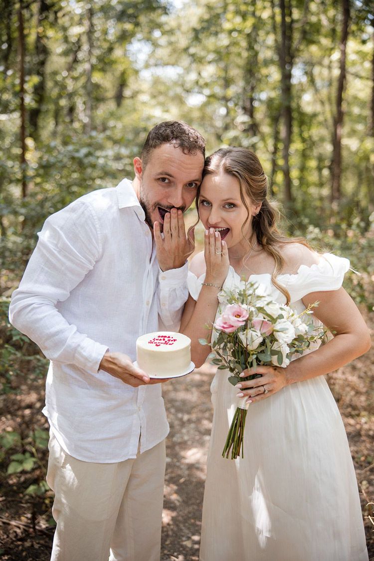 A couple in wedding attire holding a cake shares playful moments and laughter during an outdoor photoshoot in a sunlit forest.