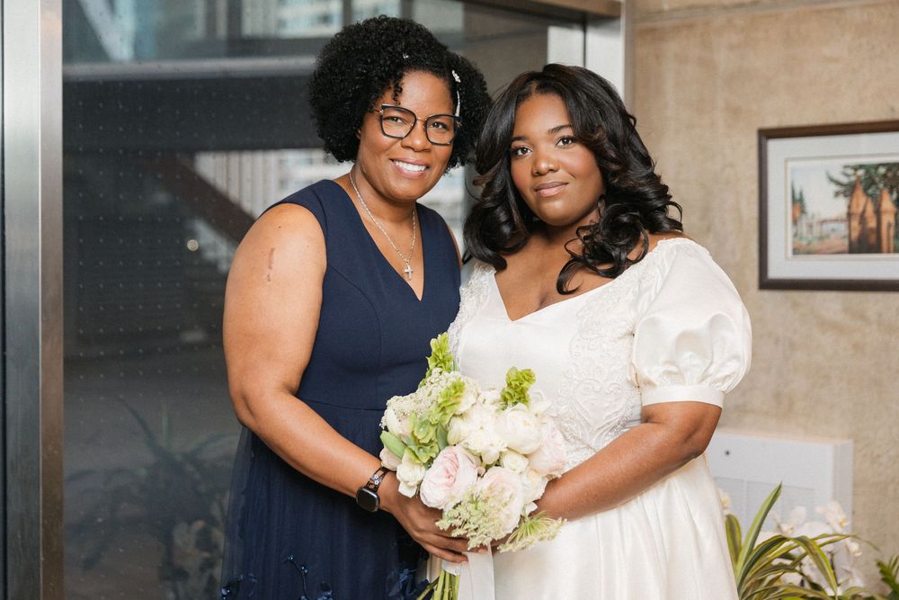 Two people pose together at a special occasion, with one holding a white and green floral bouquet.