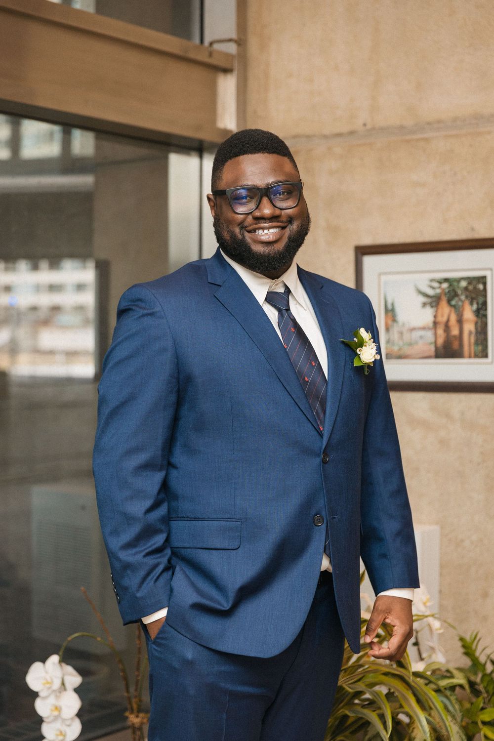 Groom in a navy blue suit with burgundy tie poses against a beige wall with indoor plants.