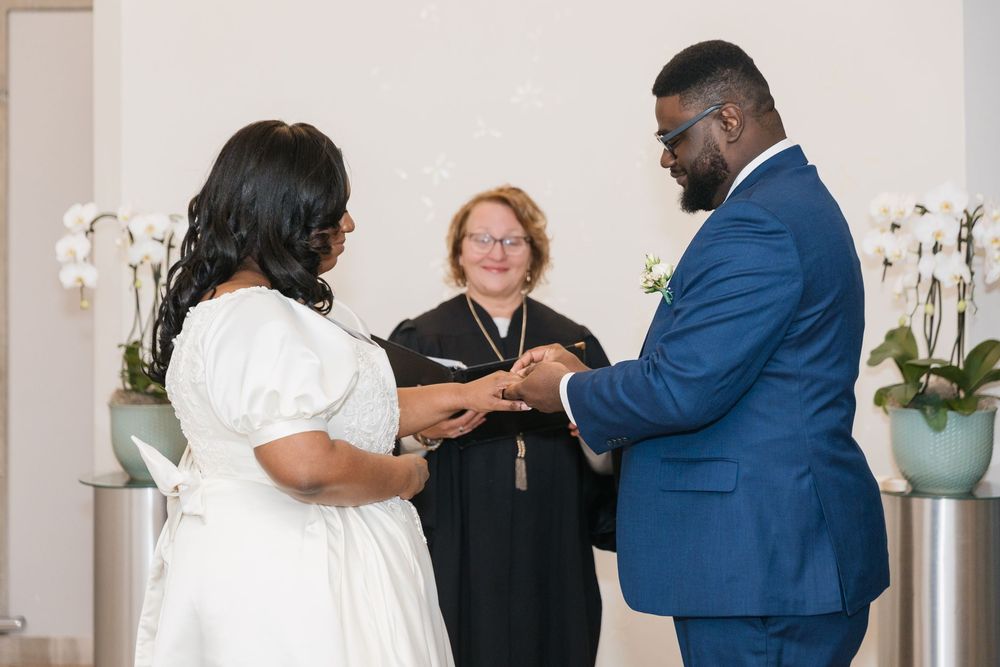A wedding ceremony takes place in an elegant white room decorated with orchids and soft lighting.