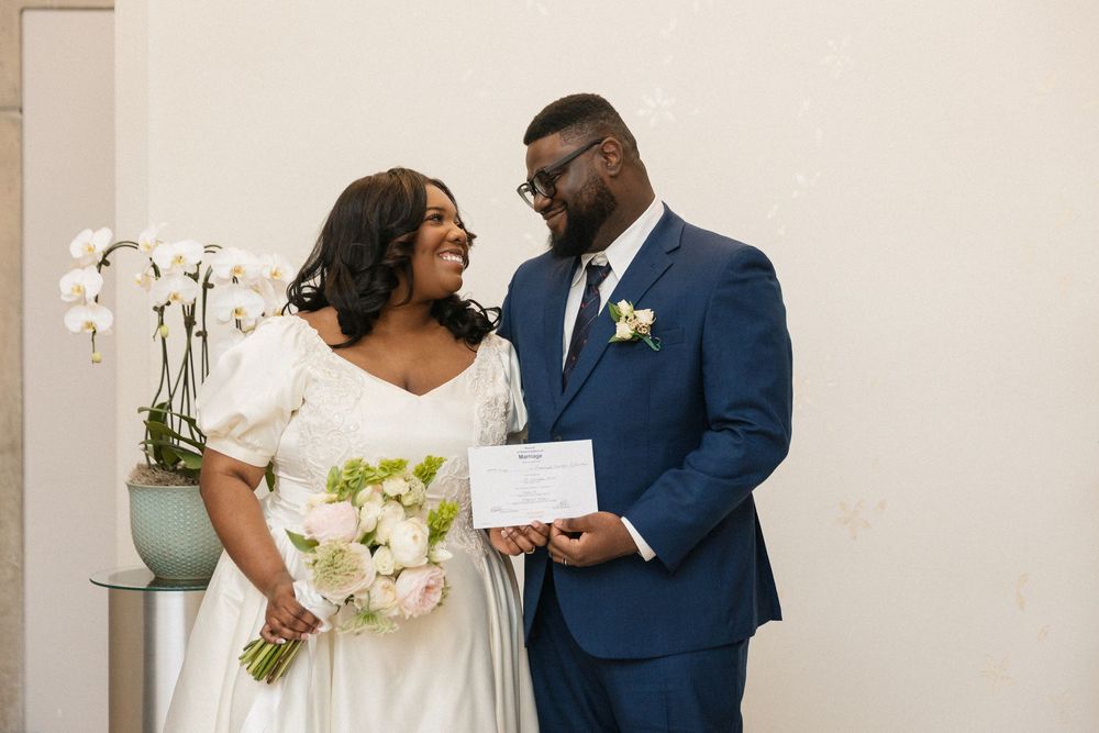A couple in wedding attire, wearing navy suit and white dress, pose together in an elegant indoor setting.